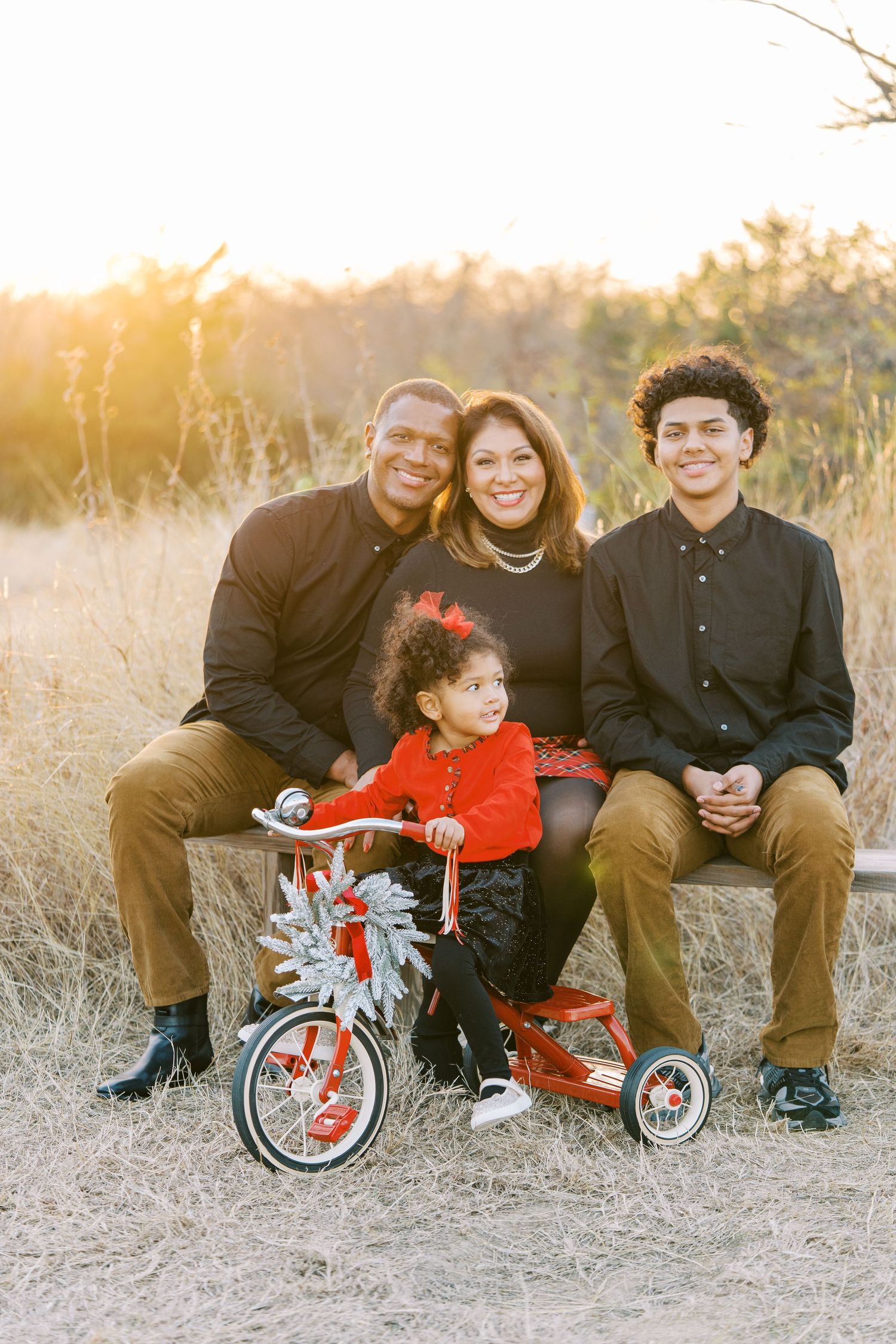 Family enjoys outdoor photo session with child on red tricycle amid golden grass at sunset.