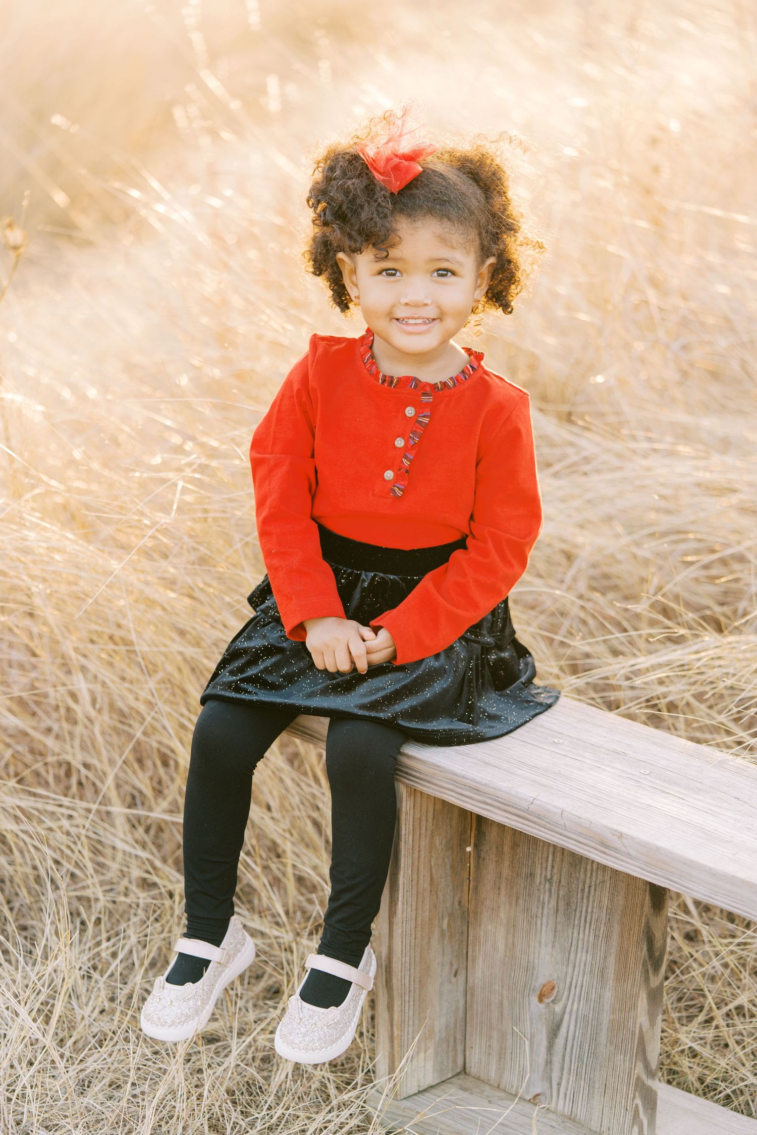 Child in bright red sweater and black pants sits happily on wooden bench surrounded by golden grass.