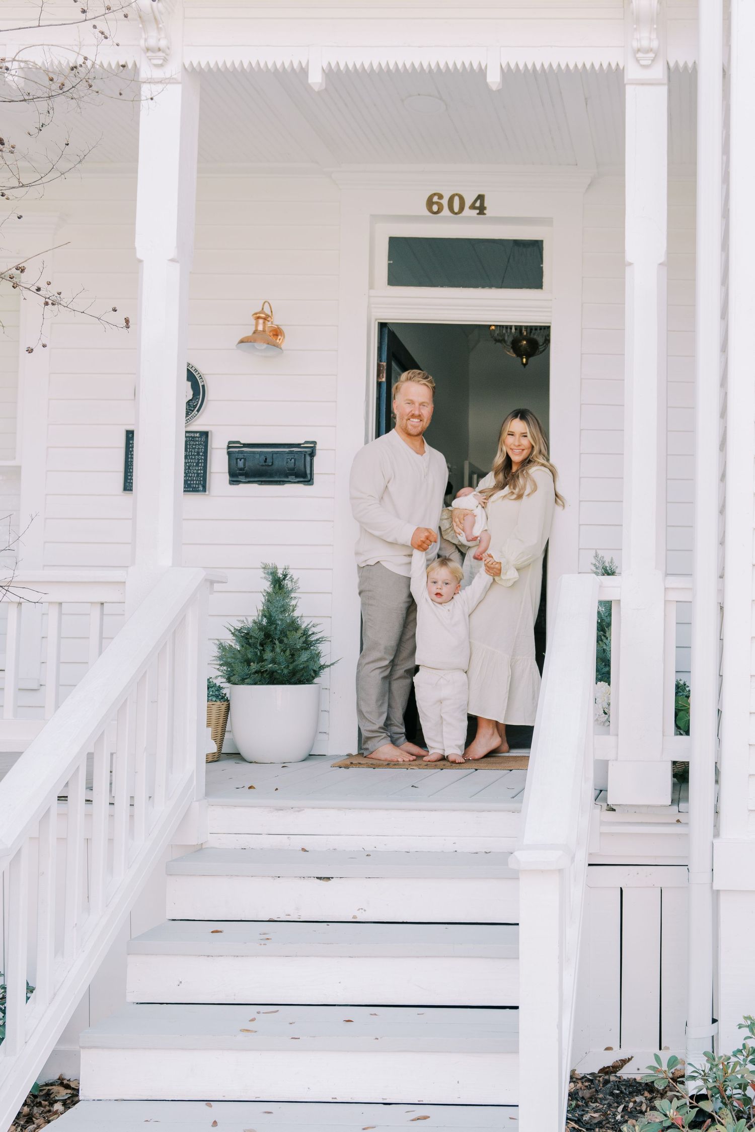 Family dressed in white poses on the porch of a classic white house with green trim.