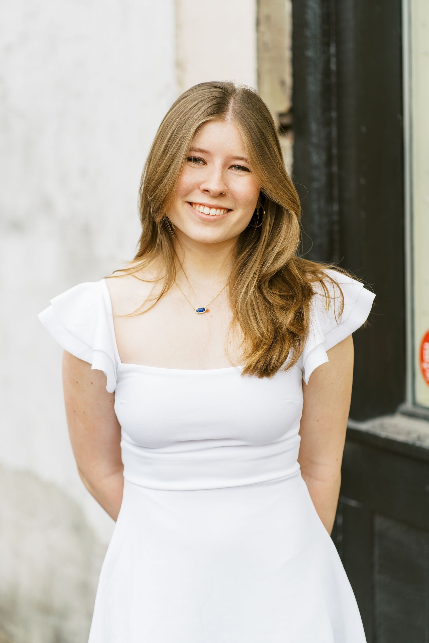 A person in a white summer dress with ruffled cap sleeves stands against a light colored wall outdoors.