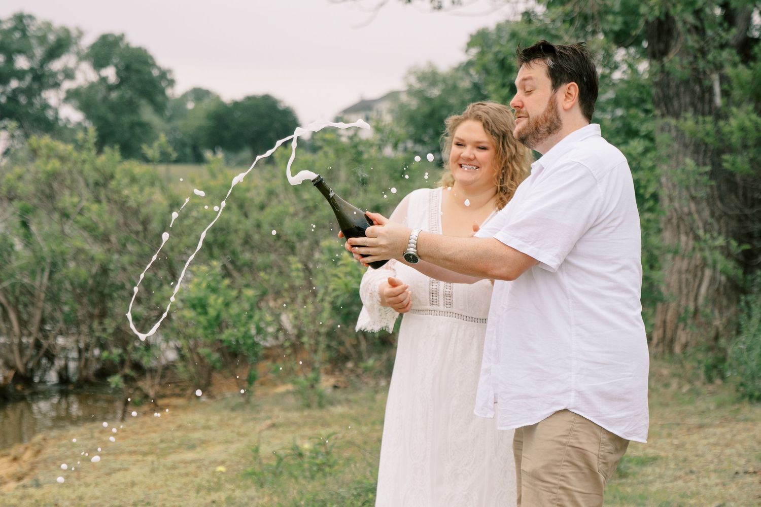 Joyful couple popping champagne bottle together during outdoor engagement photo session.
