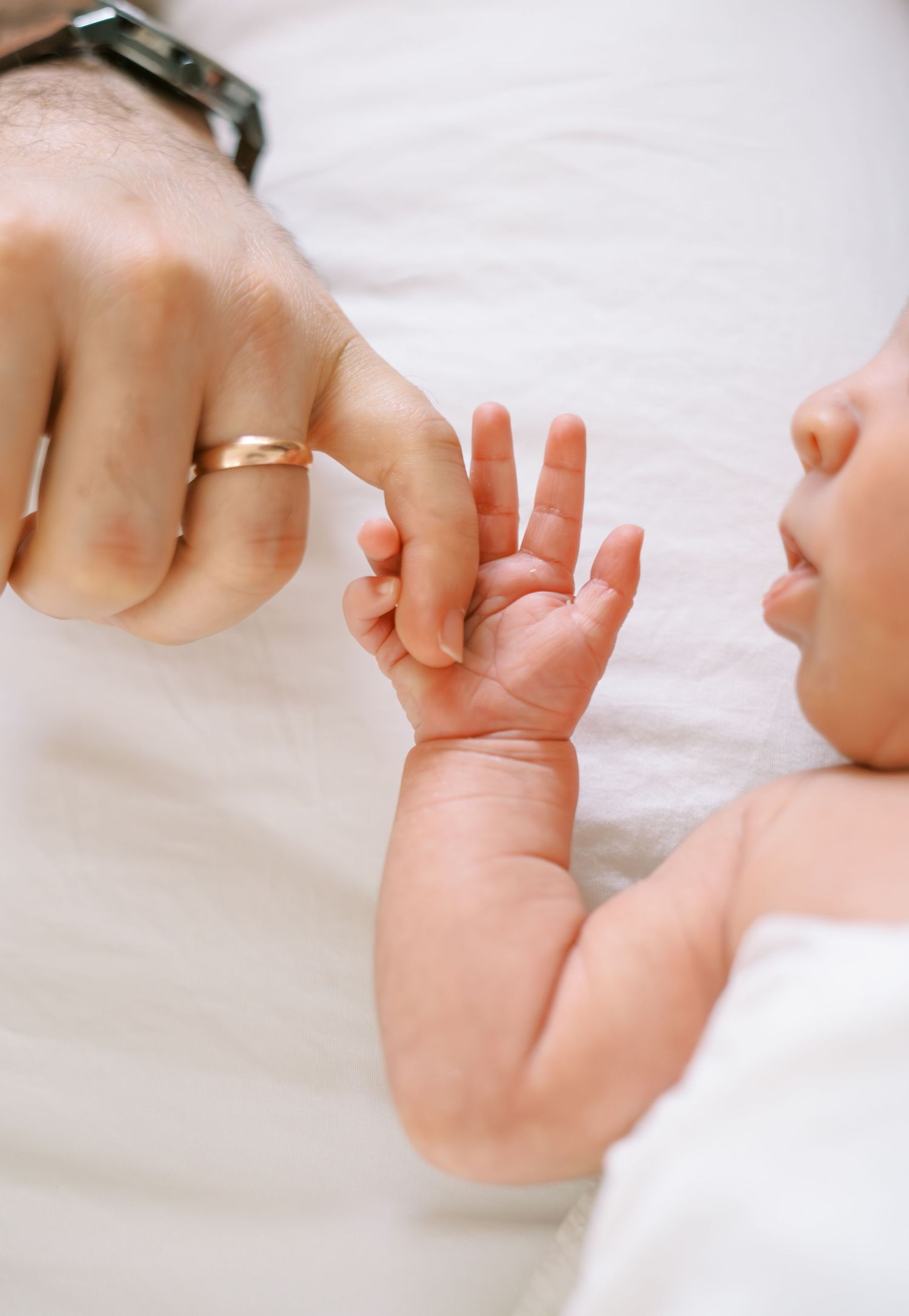 A newborn baby grasping a parent's finger, with a gold wedding ring visible on the adult's hand.