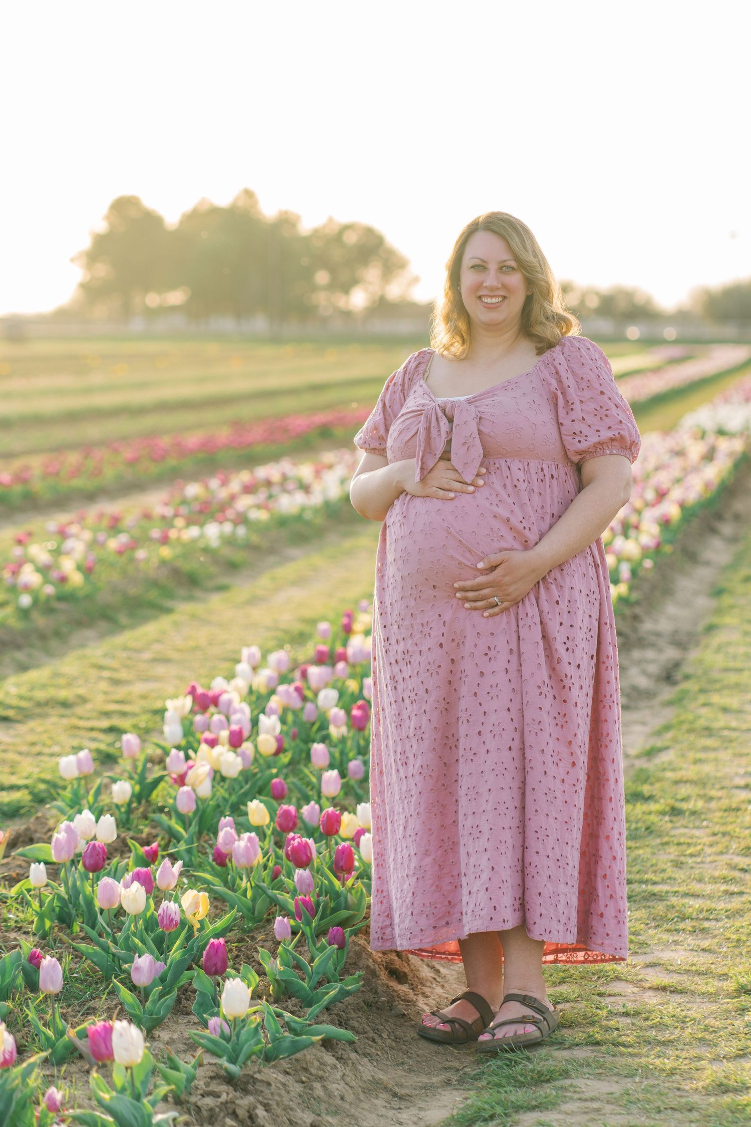 Pregnant woman in pink eyelet dress standing in colorful tulip field at golden hour, cradling baby bump.