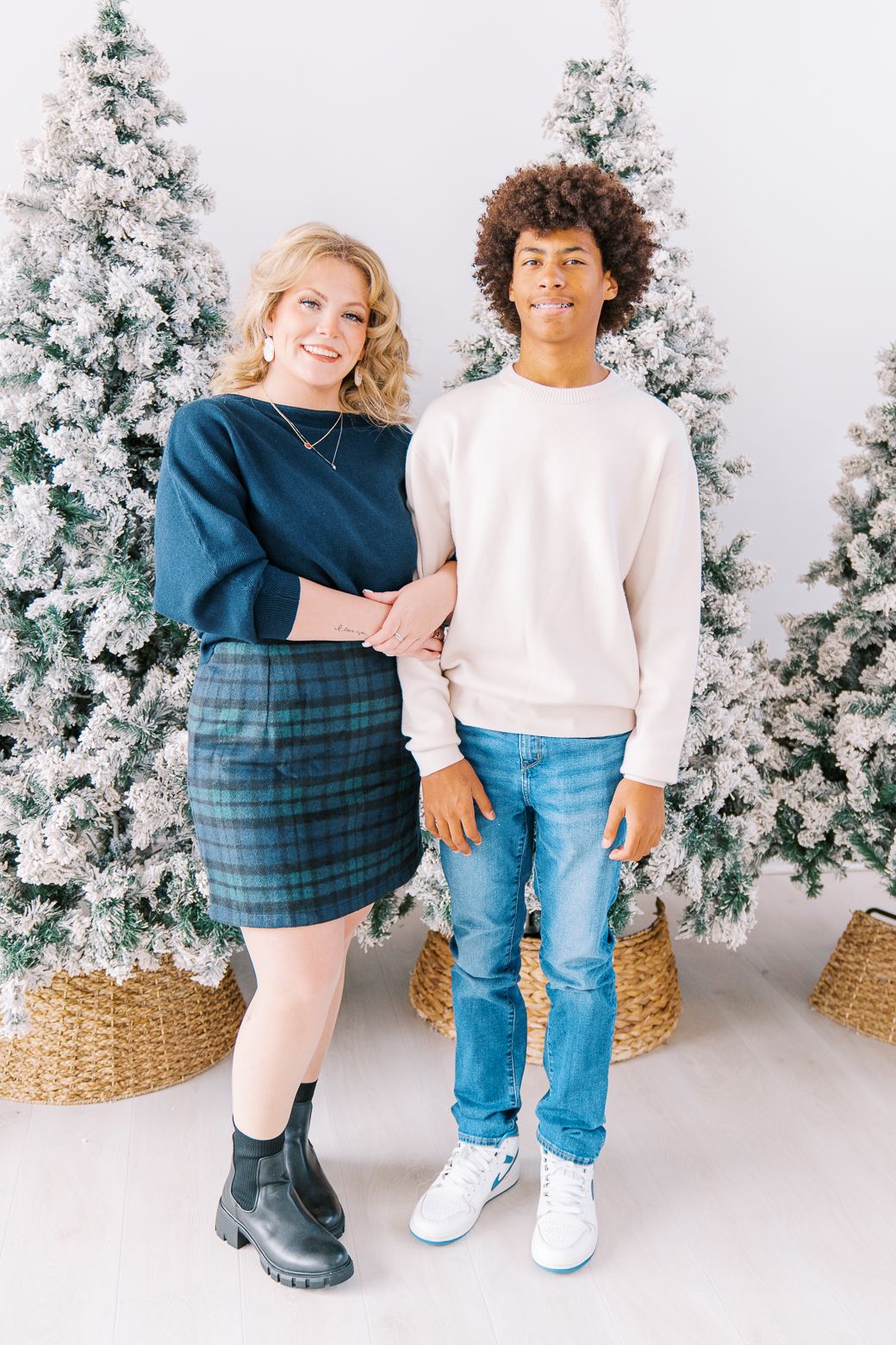 Two people pose together in front of snow-covered Christmas trees wearing casual outfits in a winter holiday photoshoot.