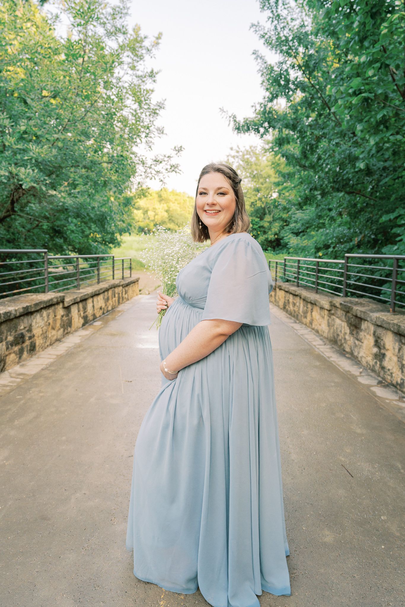 Expectant mother in flowing dusty blue gown poses on stone bridge surrounded by lush greenery.