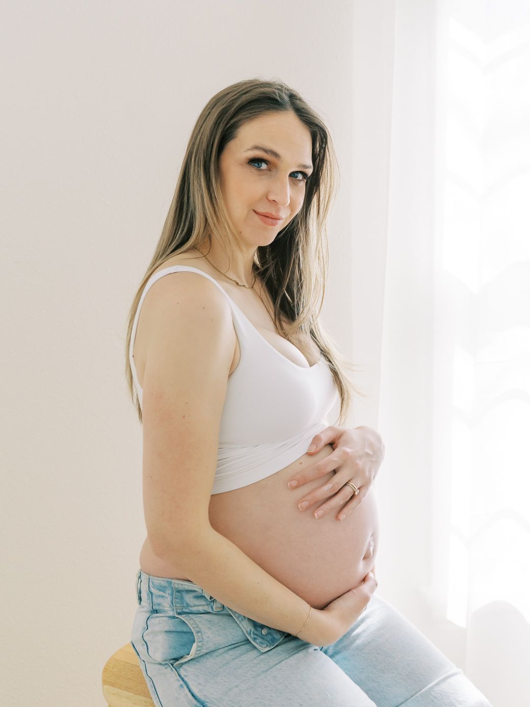 An expectant mother in light denim and white tank top poses gracefully by a bright window.
