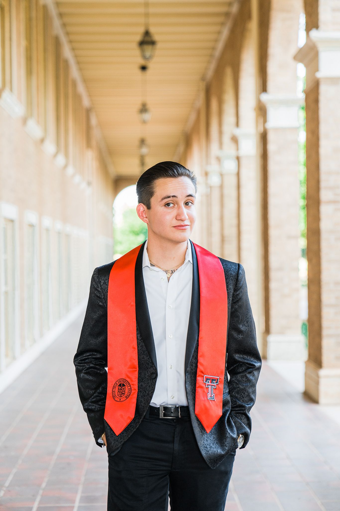 Graduate in formal attire with red graduation stole stands in front of classical architecture.