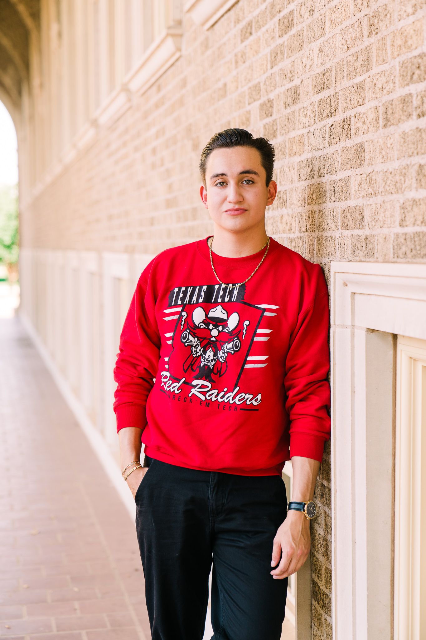 A person wearing a red Texas Tech sweatshirt and black pants stands against a brick wall.