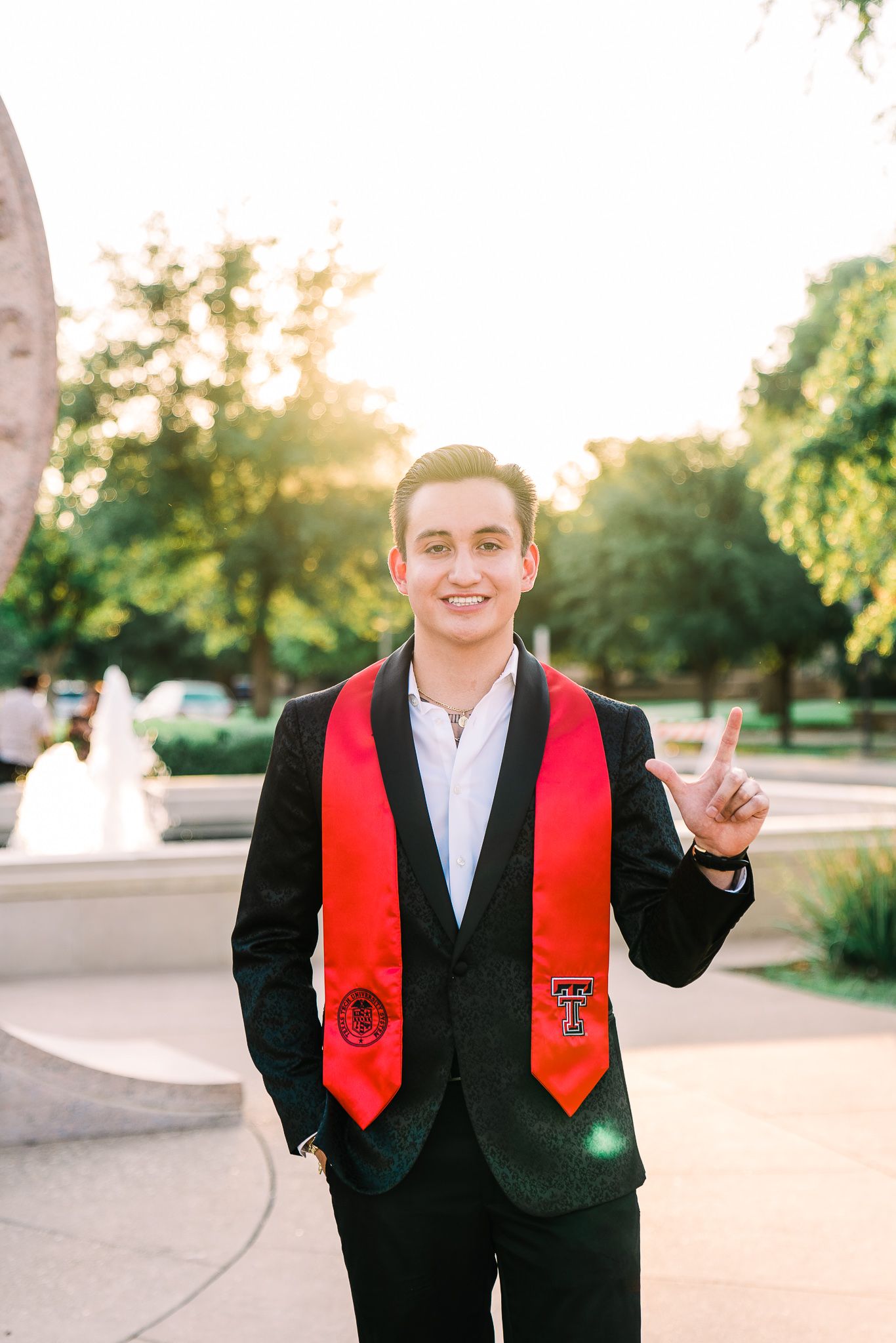 Graduate wearing black suit and red stole poses outdoors during sunset celebration.