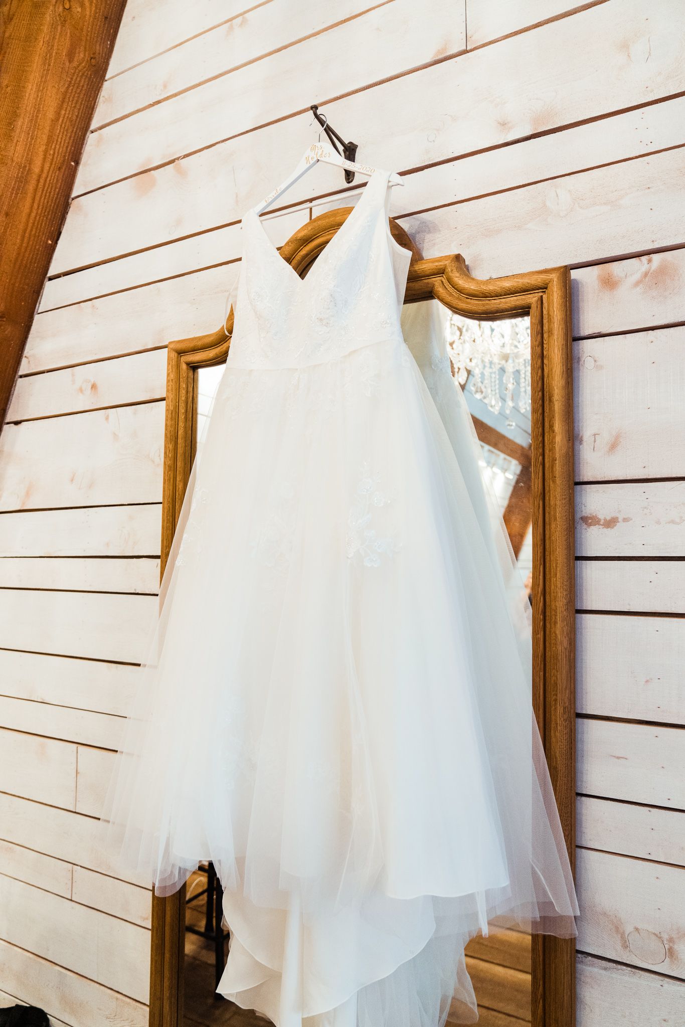 A white wedding dress with tulle skirt hangs on display against a rustic wooden wall with a vintage mirror.