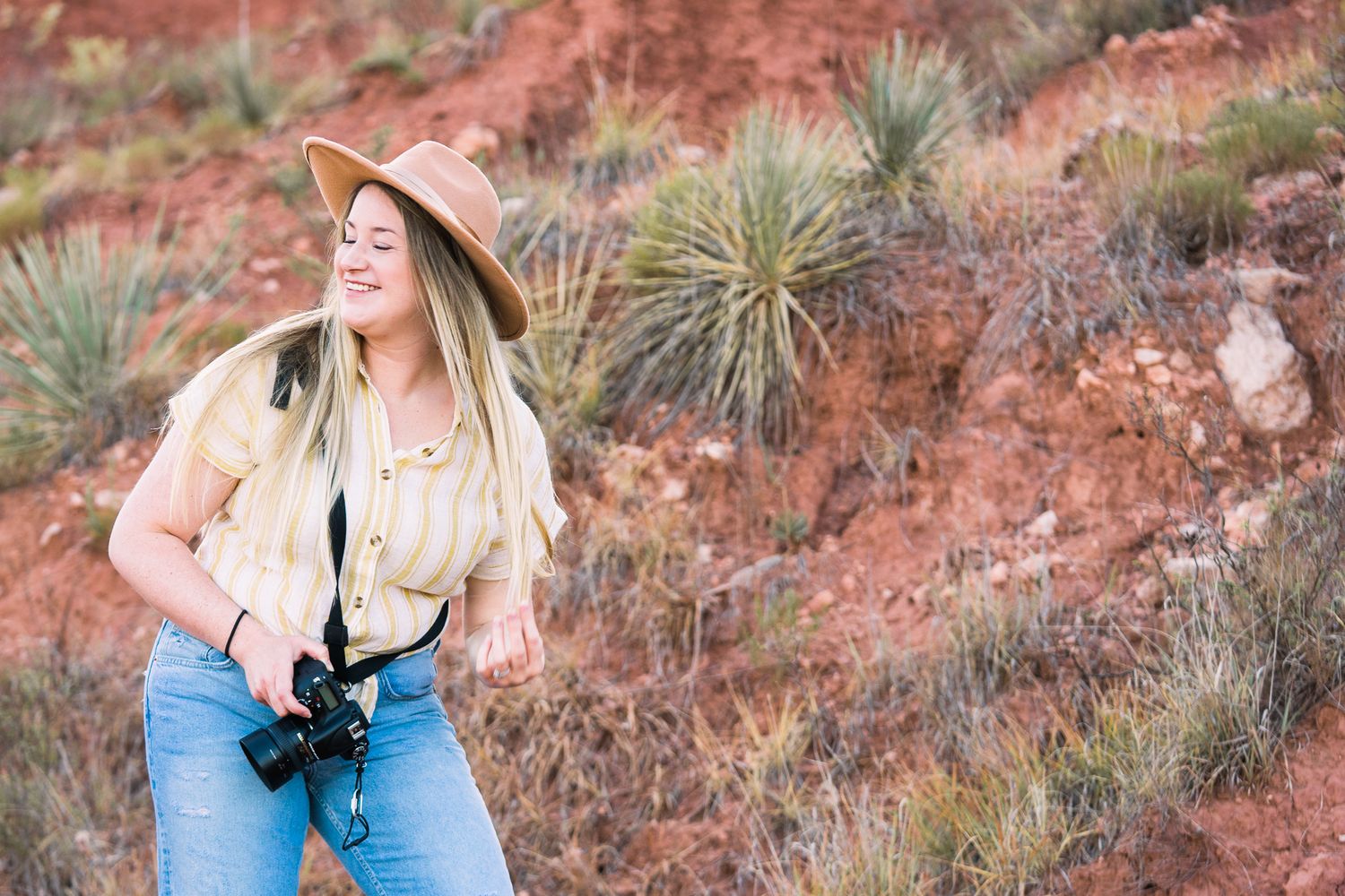 Woman in blue dress and sun hat poses against desert landscape with red rocks and desert plants in background.