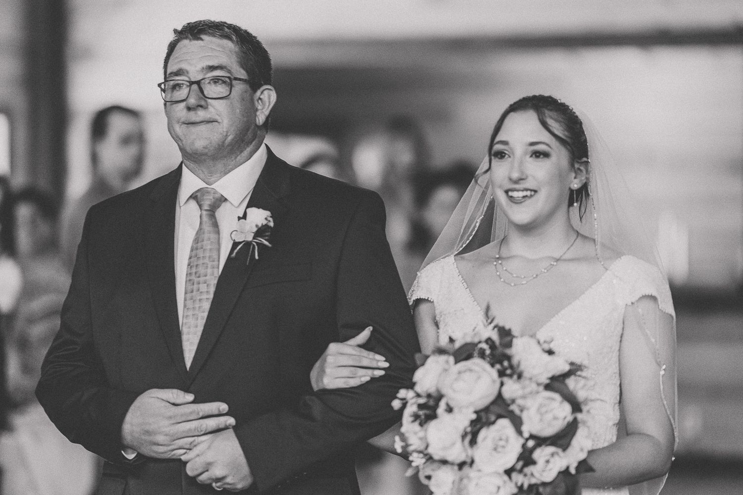 Black and white photo of a wedding ceremony with a couple walking arm in arm, bride holding white floral bouquet.