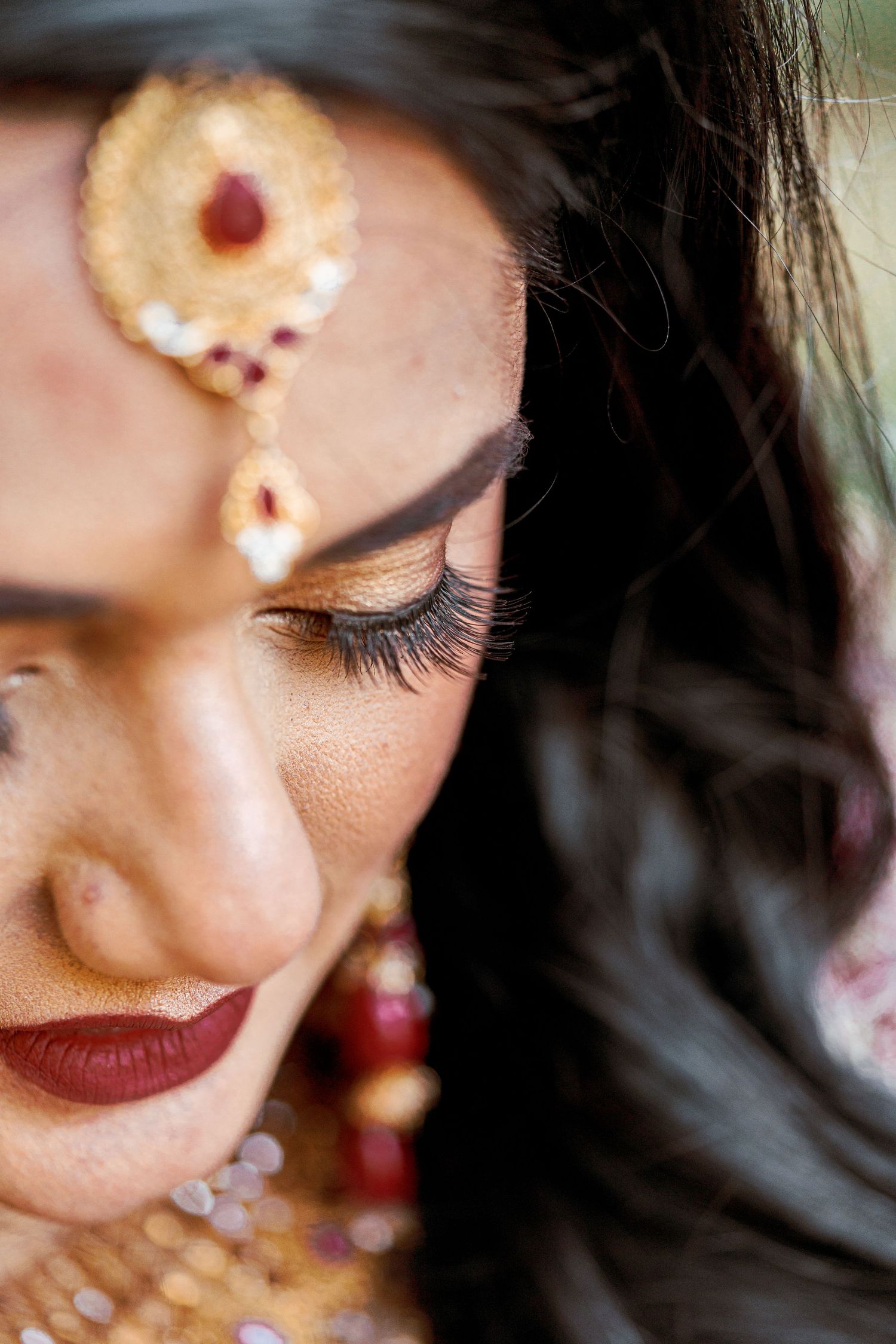 Close-up details of bridal makeup and jewelry with burgundy lipstick and gold accents.
