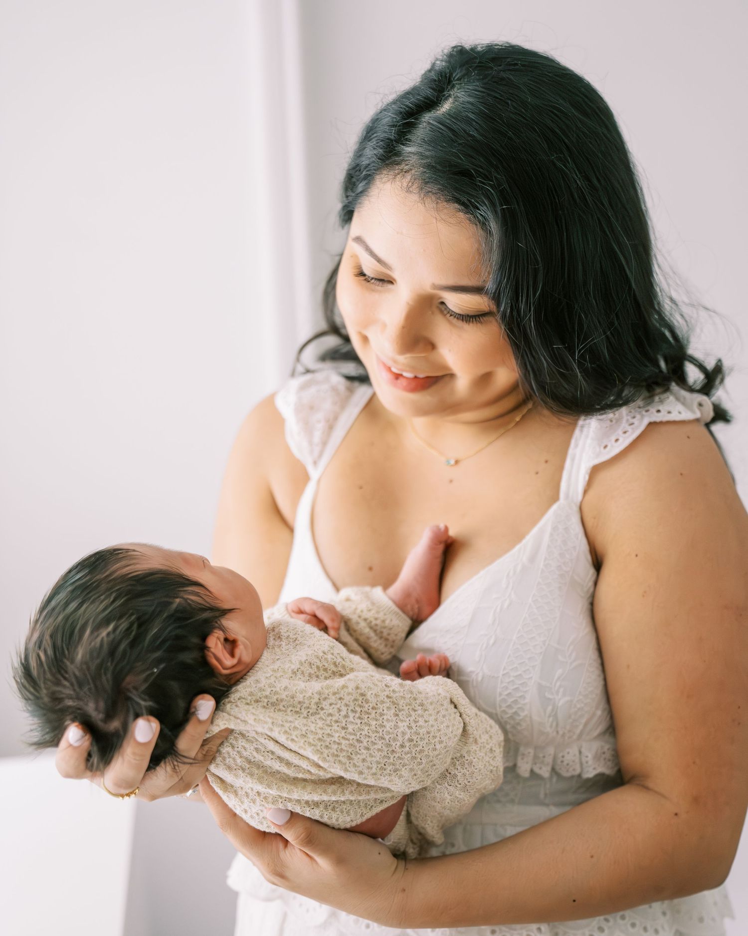 A mother tenderly holds her newborn baby, dressed in white, in a softly lit studio setting.