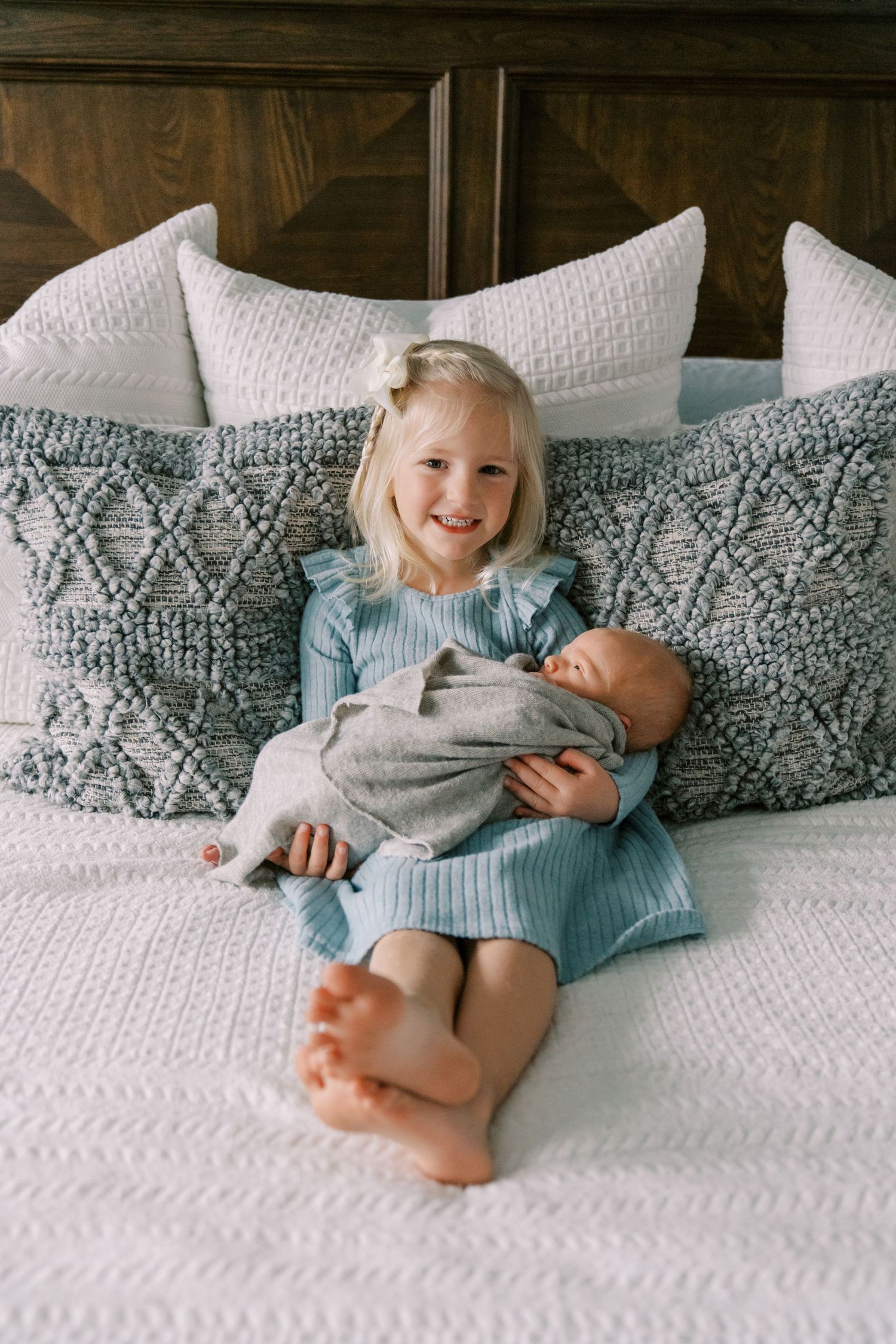 Young girl in blue dress smiles while holding newborn baby sibling on a white bed with gray pillows.