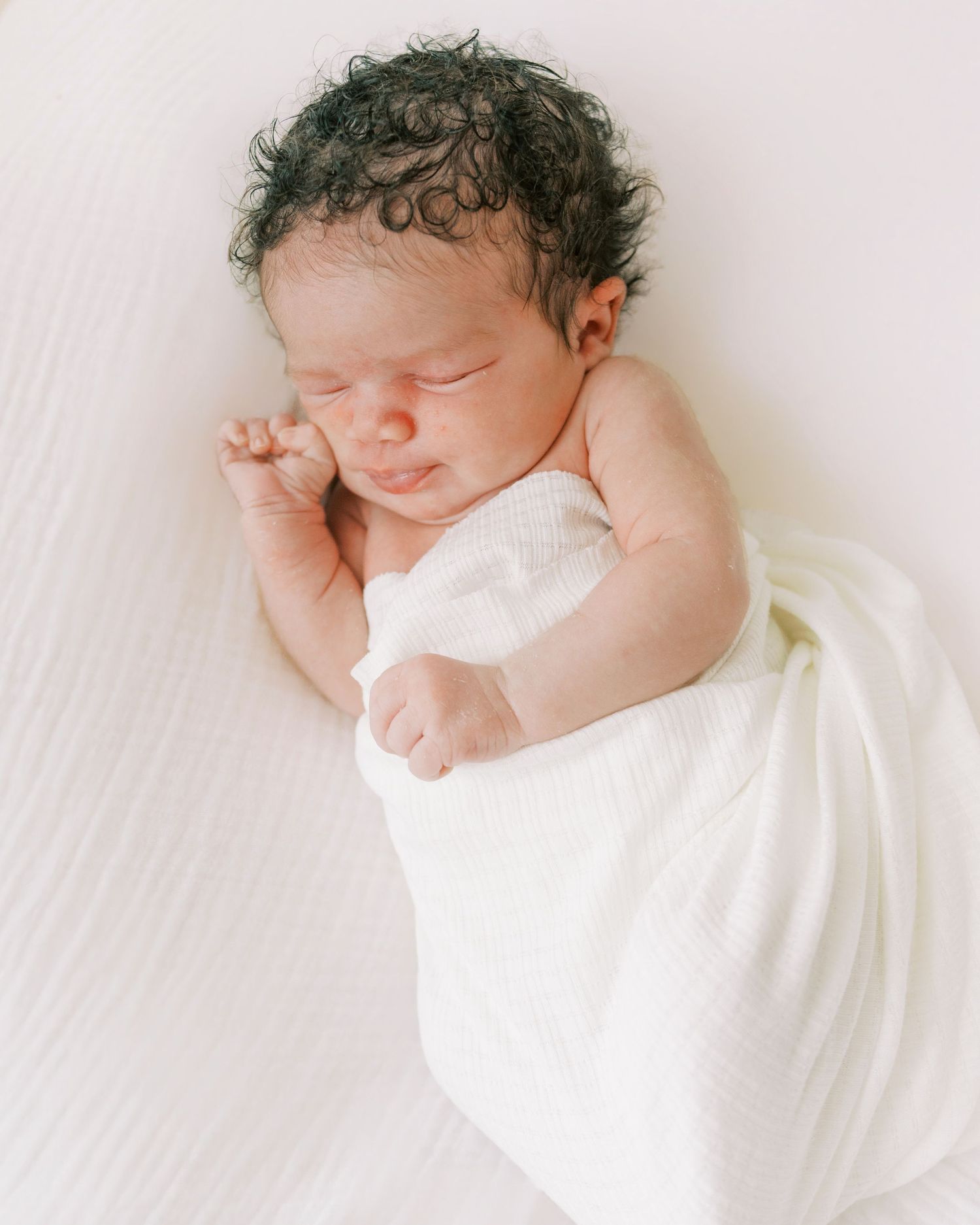 Peaceful newborn baby with dark curly hair sleeping wrapped in a soft white swaddle blanket.
