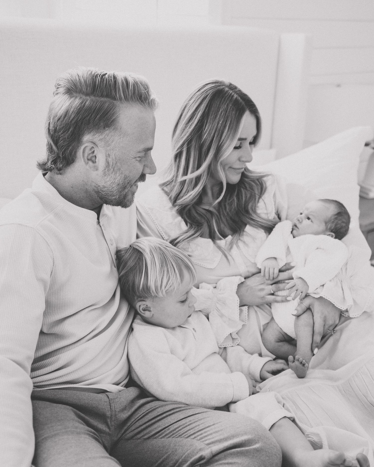 Black and white family photo of parents with toddler and newborn baby sitting together on a couch.