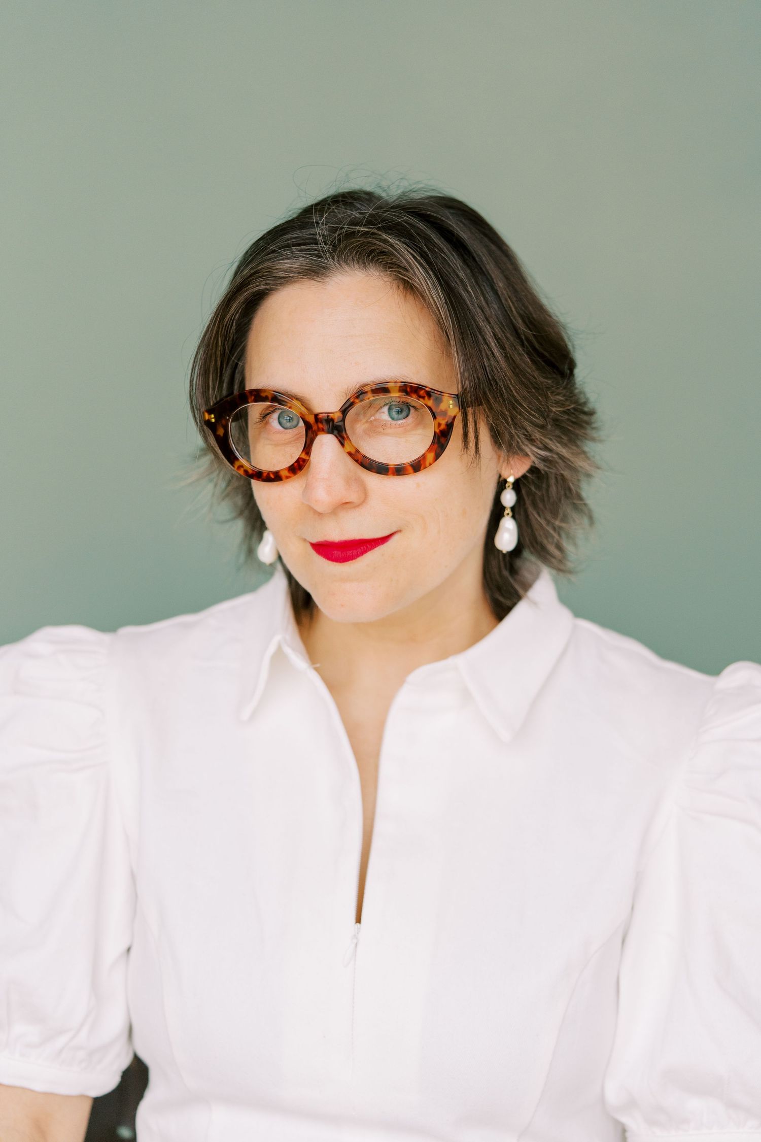 Professional portrait featuring tortoise shell glasses, pearl earrings, and a crisp white blouse against a sage green background.