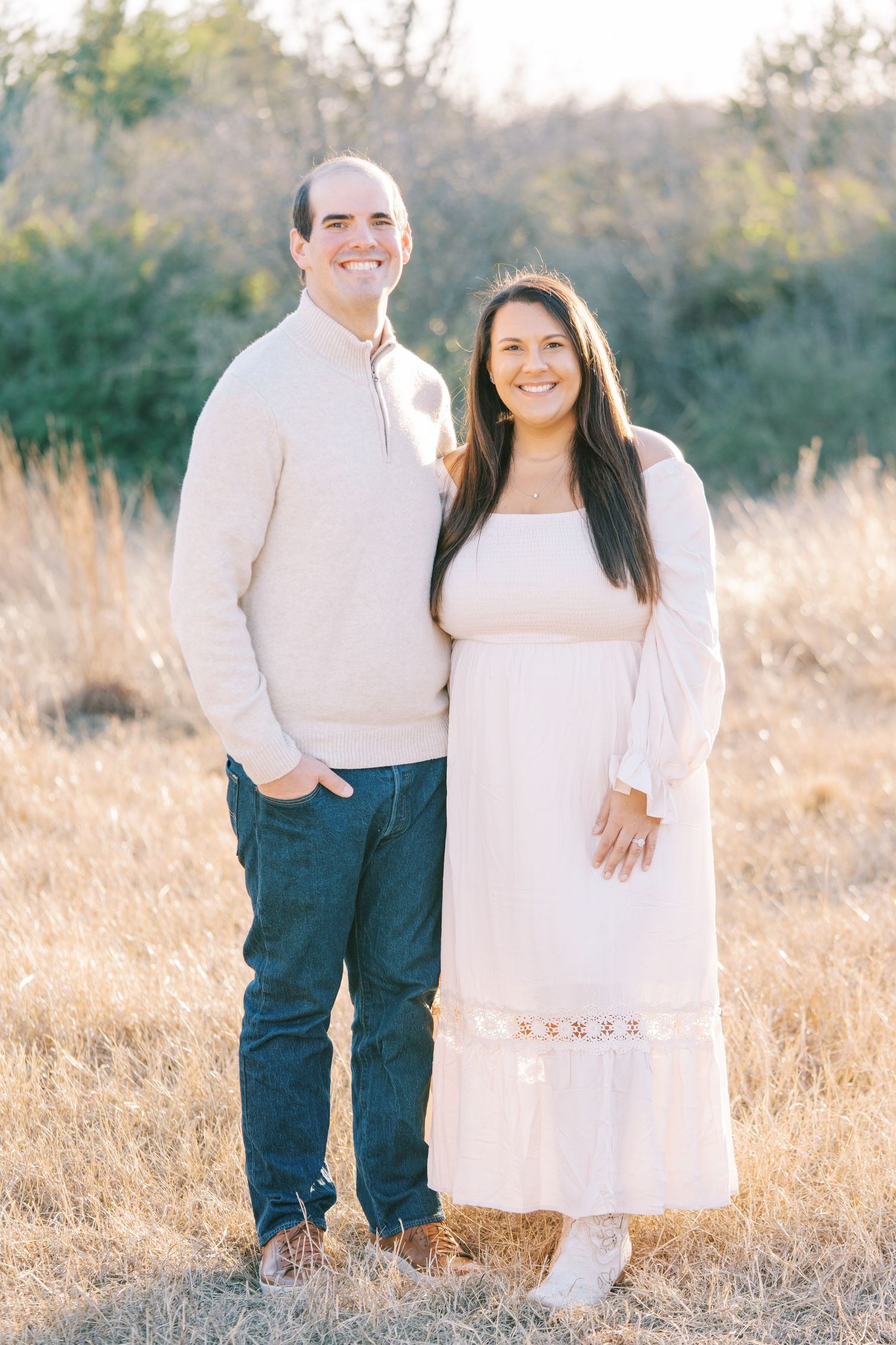 Couple in matching cream-colored outfits pose together in a golden field at sunset.