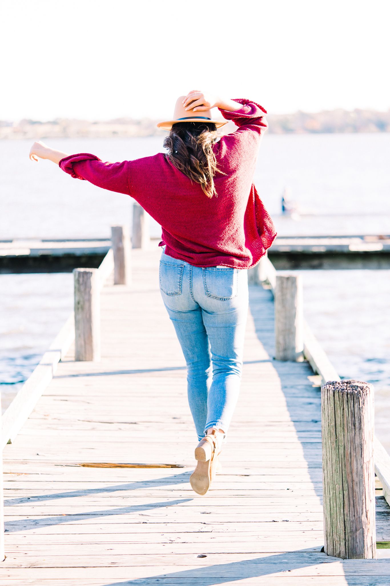 Person in red sweater and jeans jumps playfully along a wooden pier by the water on a sunny day.