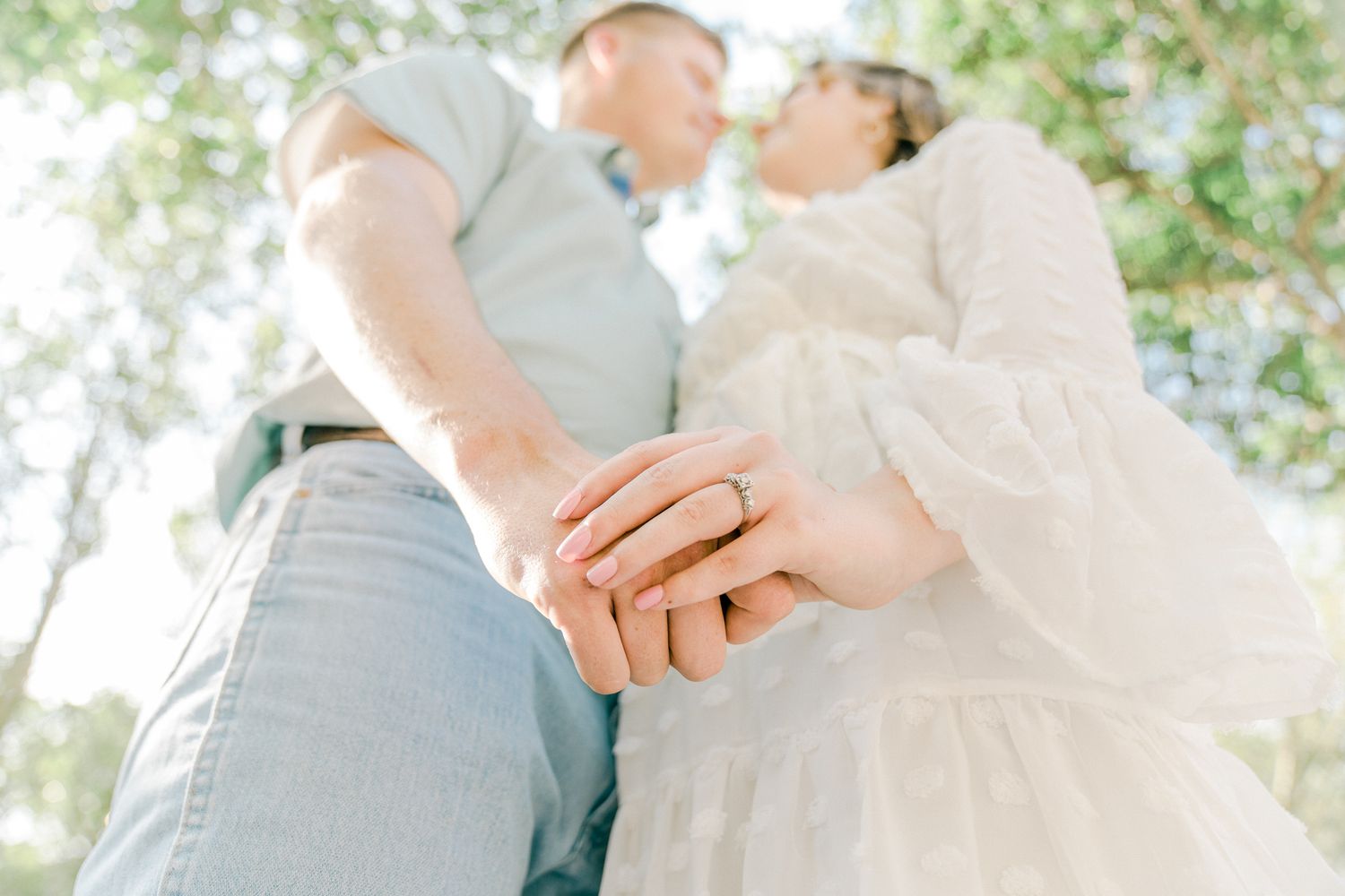 A couple holds hands while embracing outdoors with soft sunlight filtering through trees in the background.