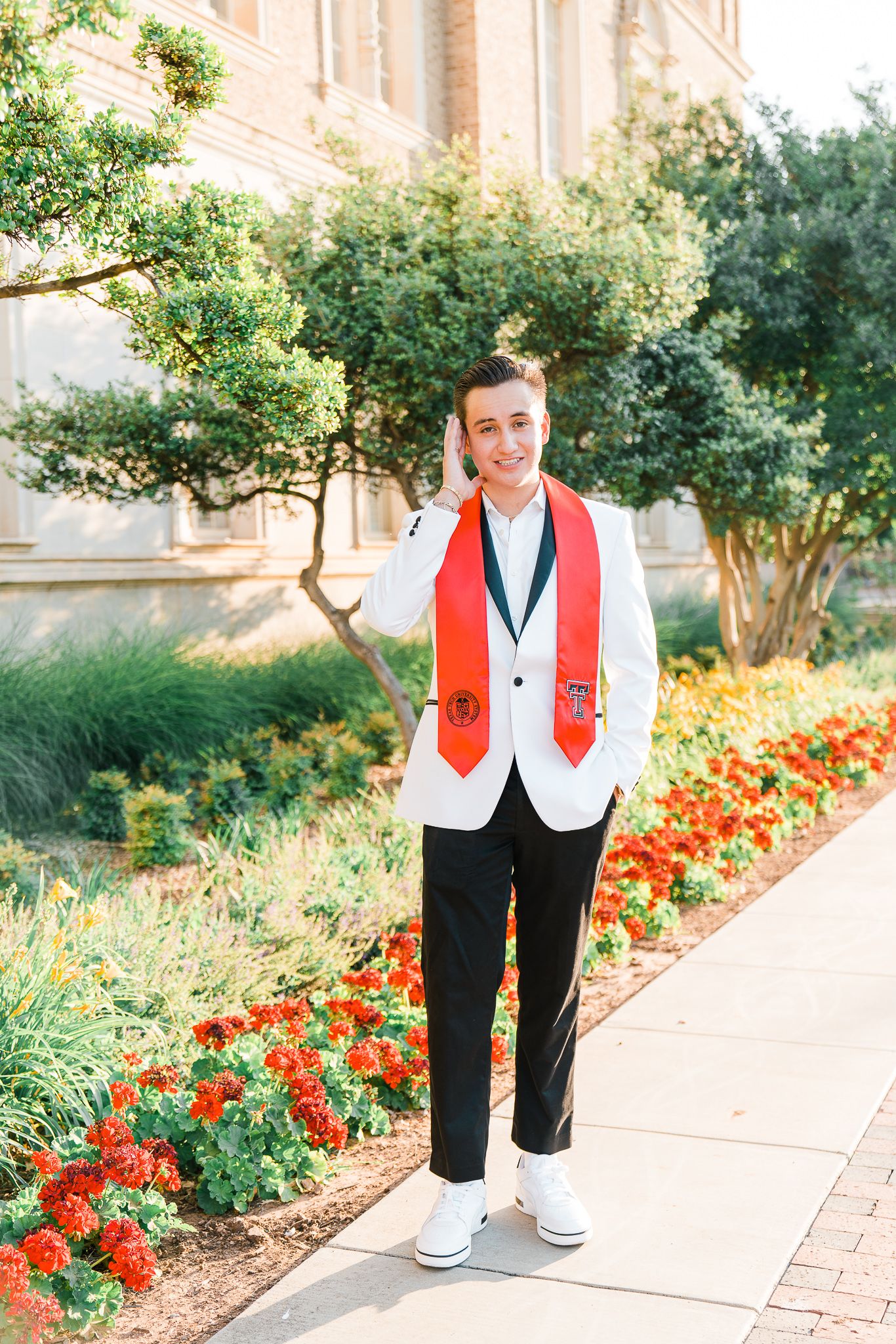 A figure in a red and white vest and black pants stands along a garden path bordered by vibrant red flowers.