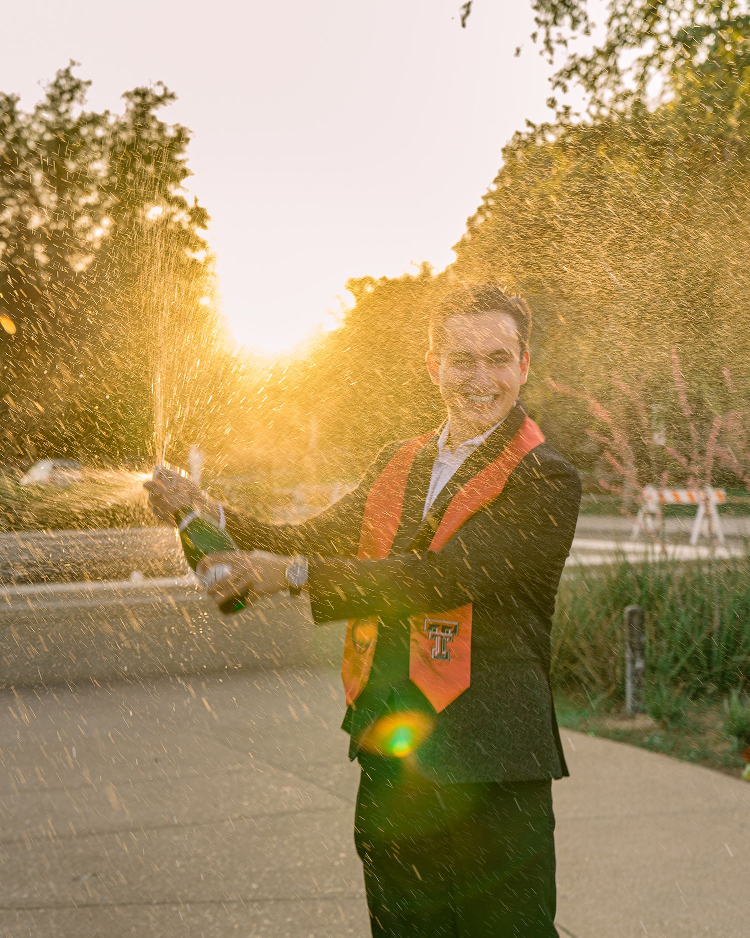 Person in dark clothing poses joyfully in a park setting with warm sunlight filtering through trees.