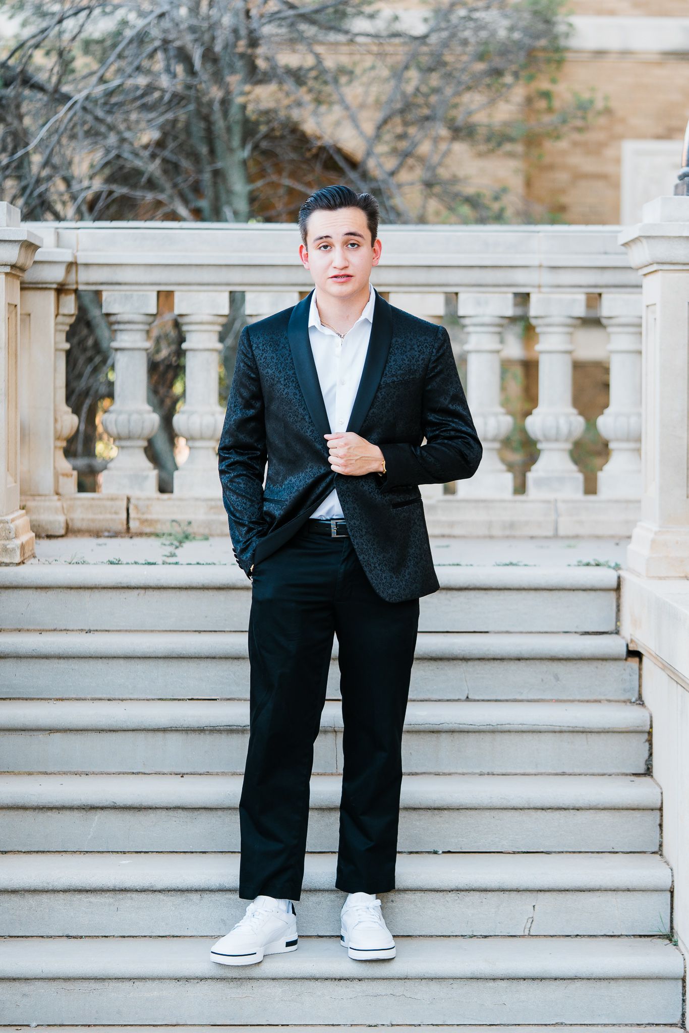 A person in a black suit and white sneakers poses casually on stone steps with decorative balustrades.