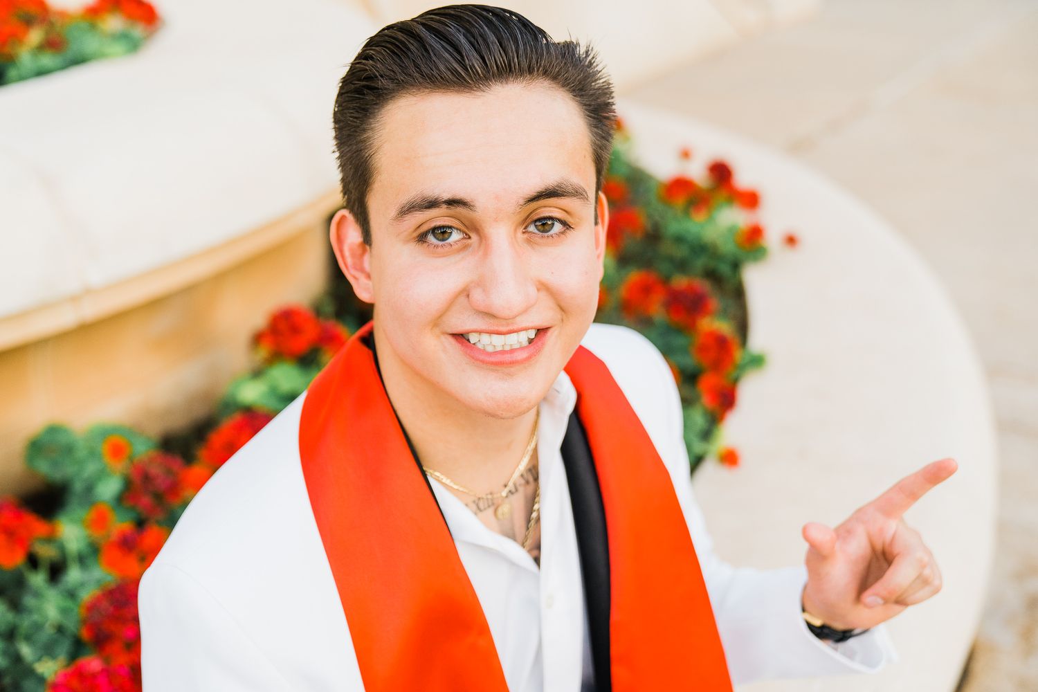 Graduate wearing red and white graduation stole smiles while pointing against floral background.