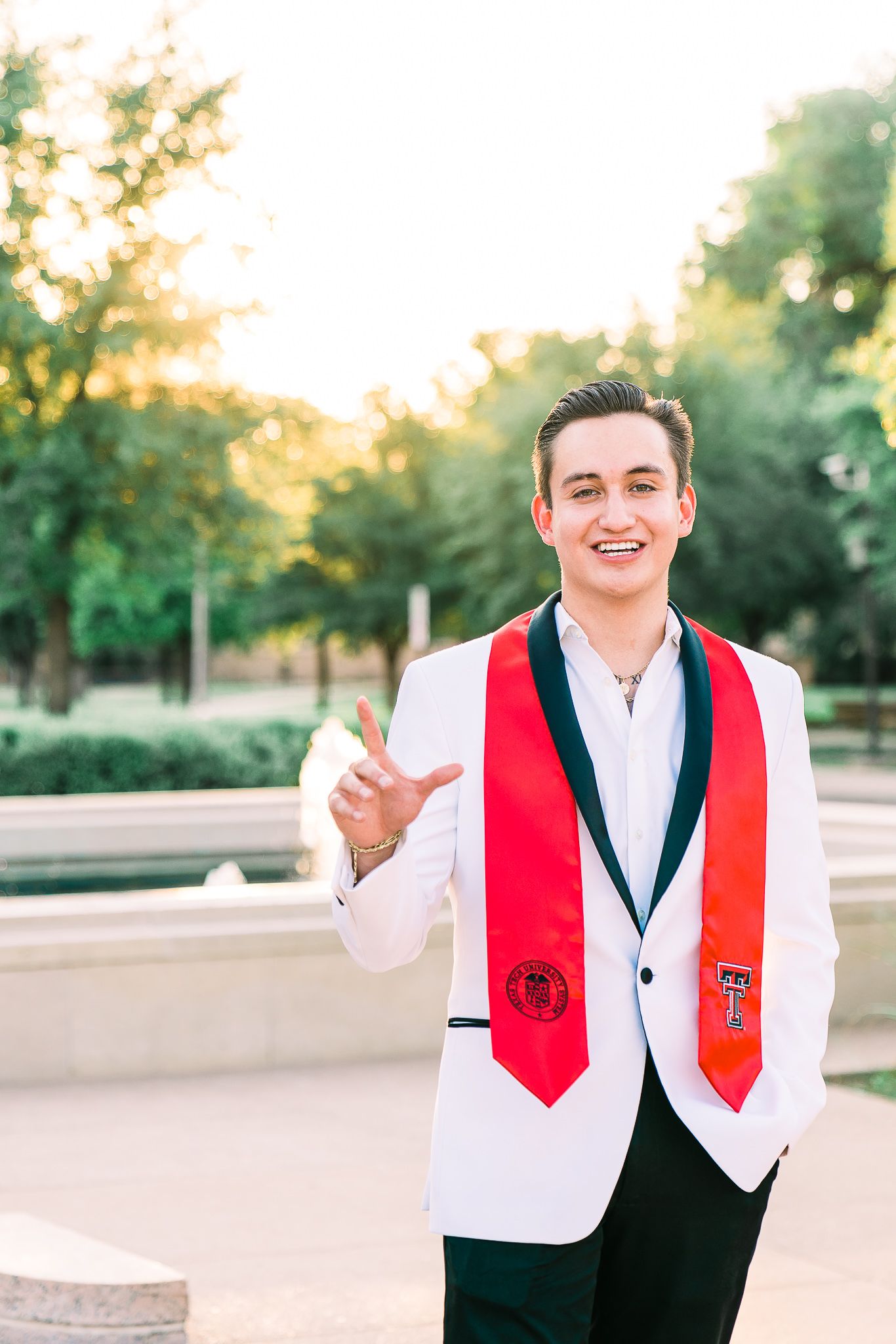 Graduate in white shirt and black vest with red stole poses outdoors during golden hour celebration.