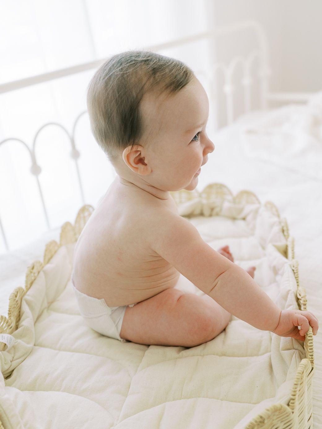 Baby resting on white bedding while looking to the side in natural light.