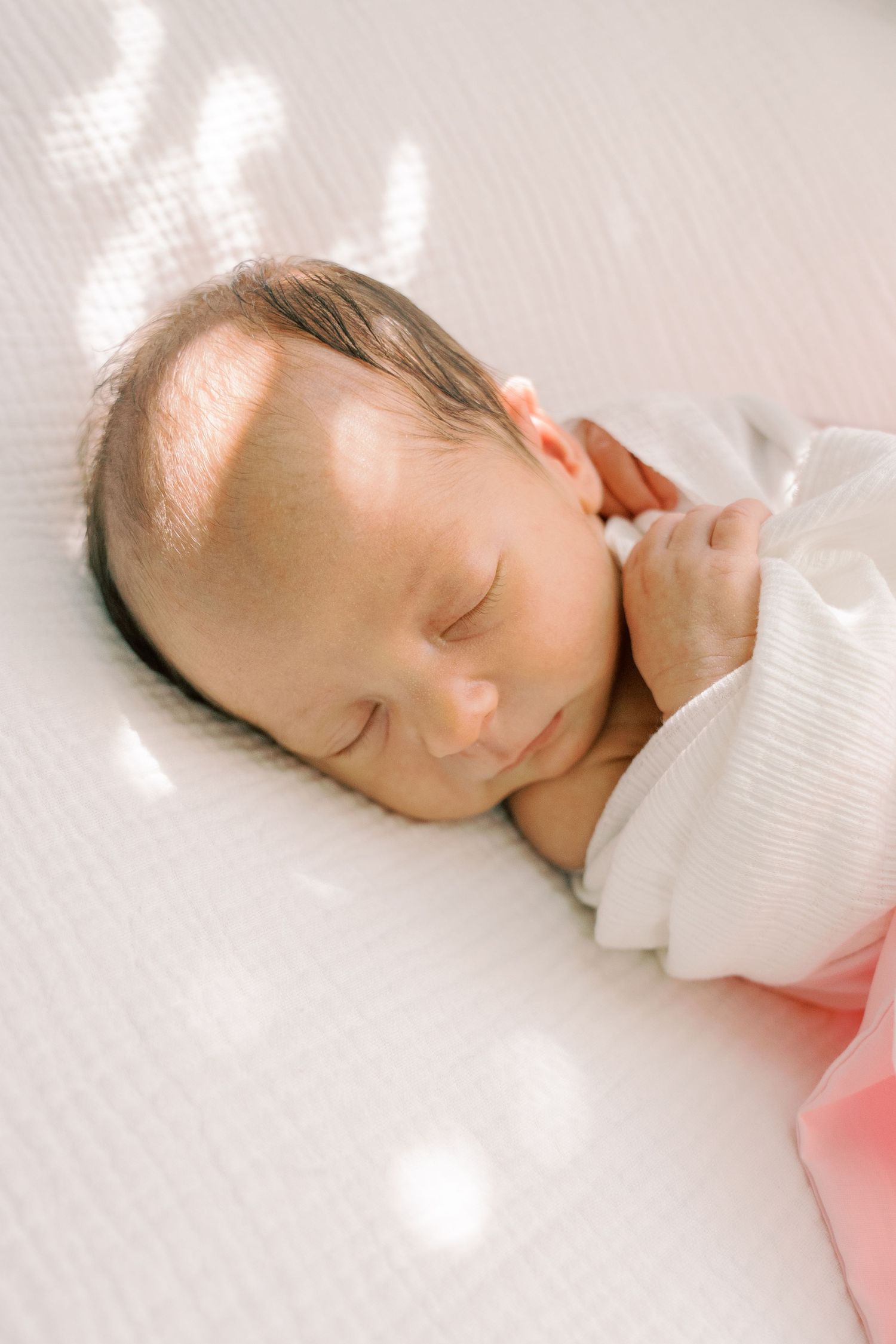 Sleeping newborn baby in soft white bedding with gentle sunlight streaming across their face.