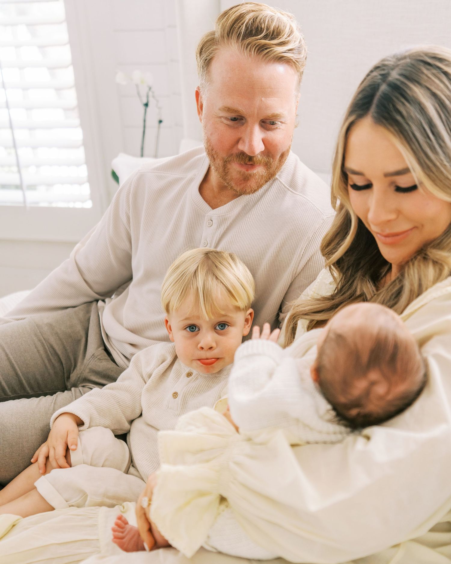 A family shares intimate moments together on a white bed in matching neutral outfits.