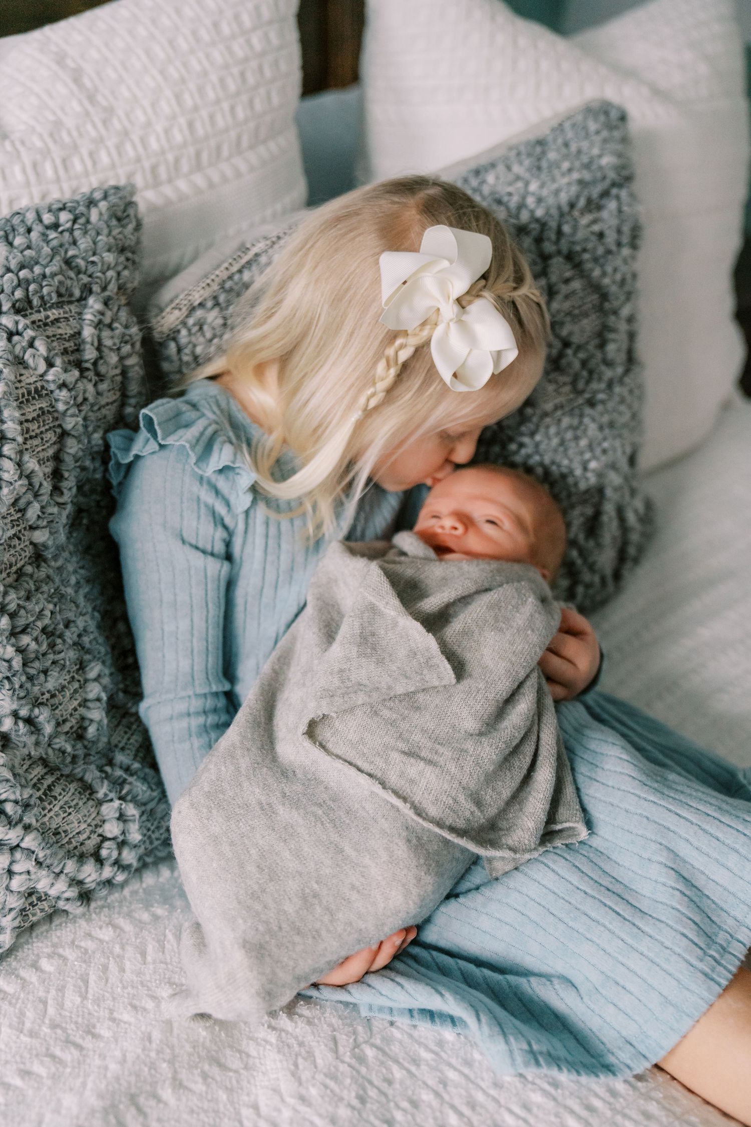 A tender moment between siblings as a young child in blue holds a swaddled newborn on a cozy bed with gray pillows.