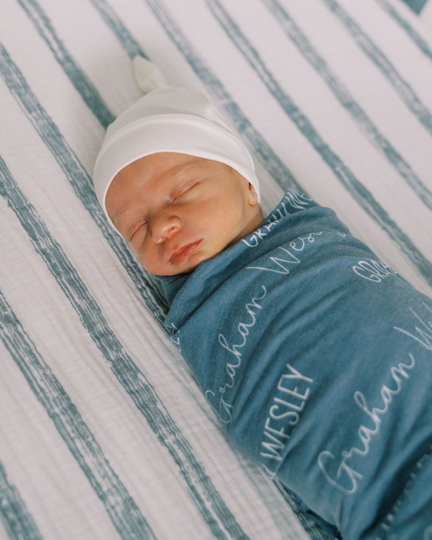 Sleeping newborn swaddled in blue text-patterned blanket on striped bedding.