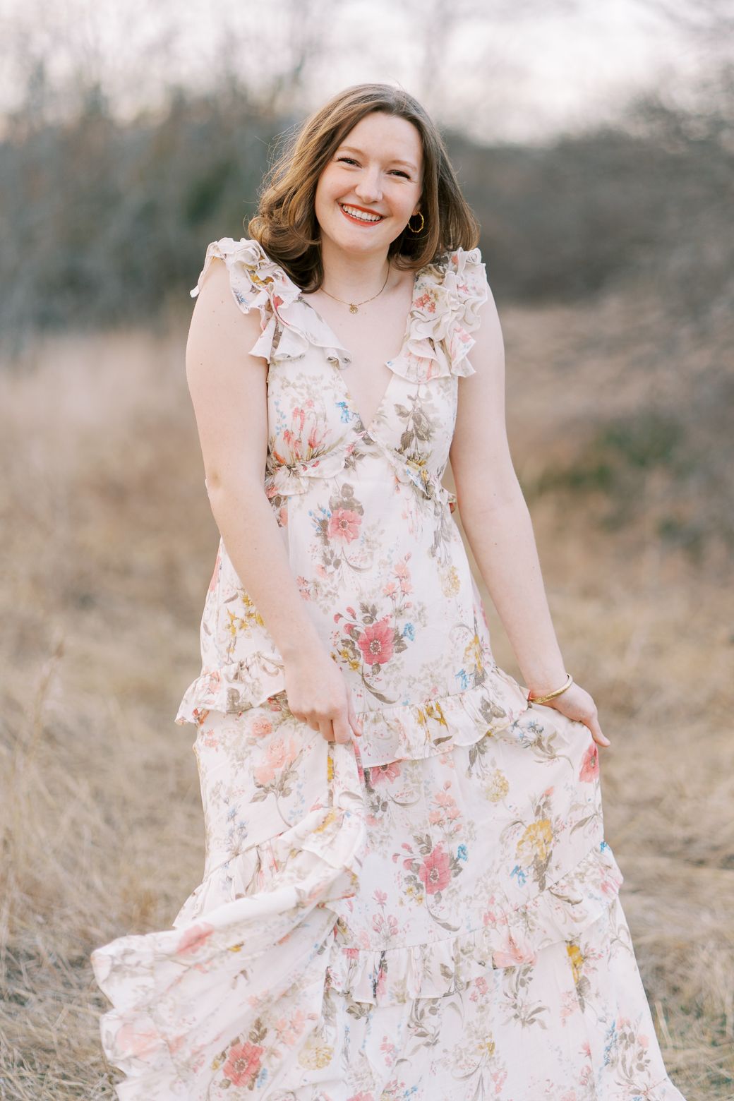 A floral dress with ruffles and pink roses is worn in a dreamy outdoor portrait session in a field.