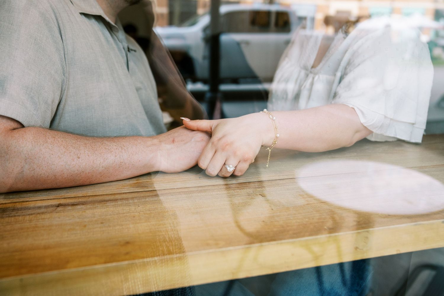 Couple holding hands across a wooden table, photographed through a window reflection.