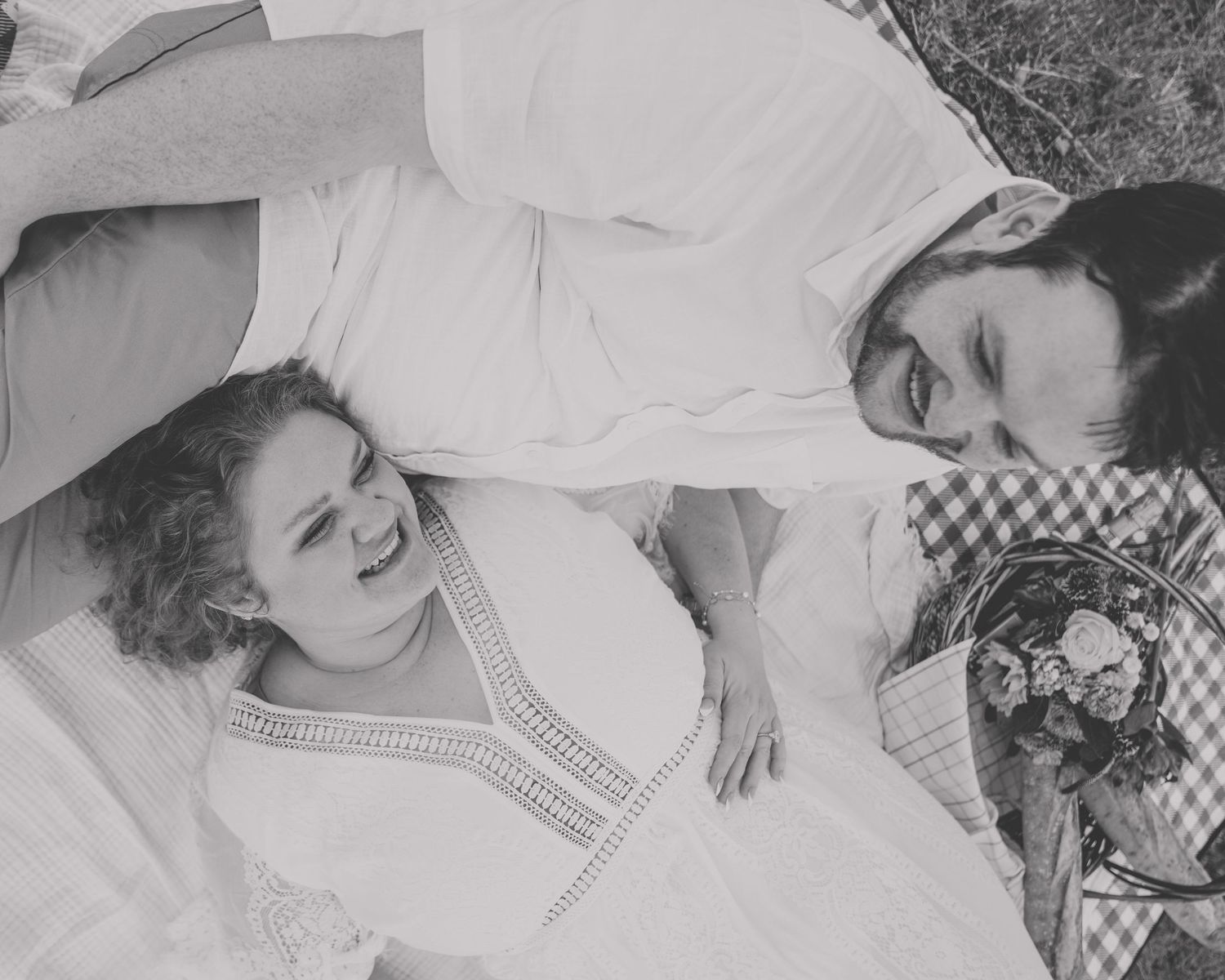 Black and white photo of couple lying on a picnic blanket laughing together outdoors.
