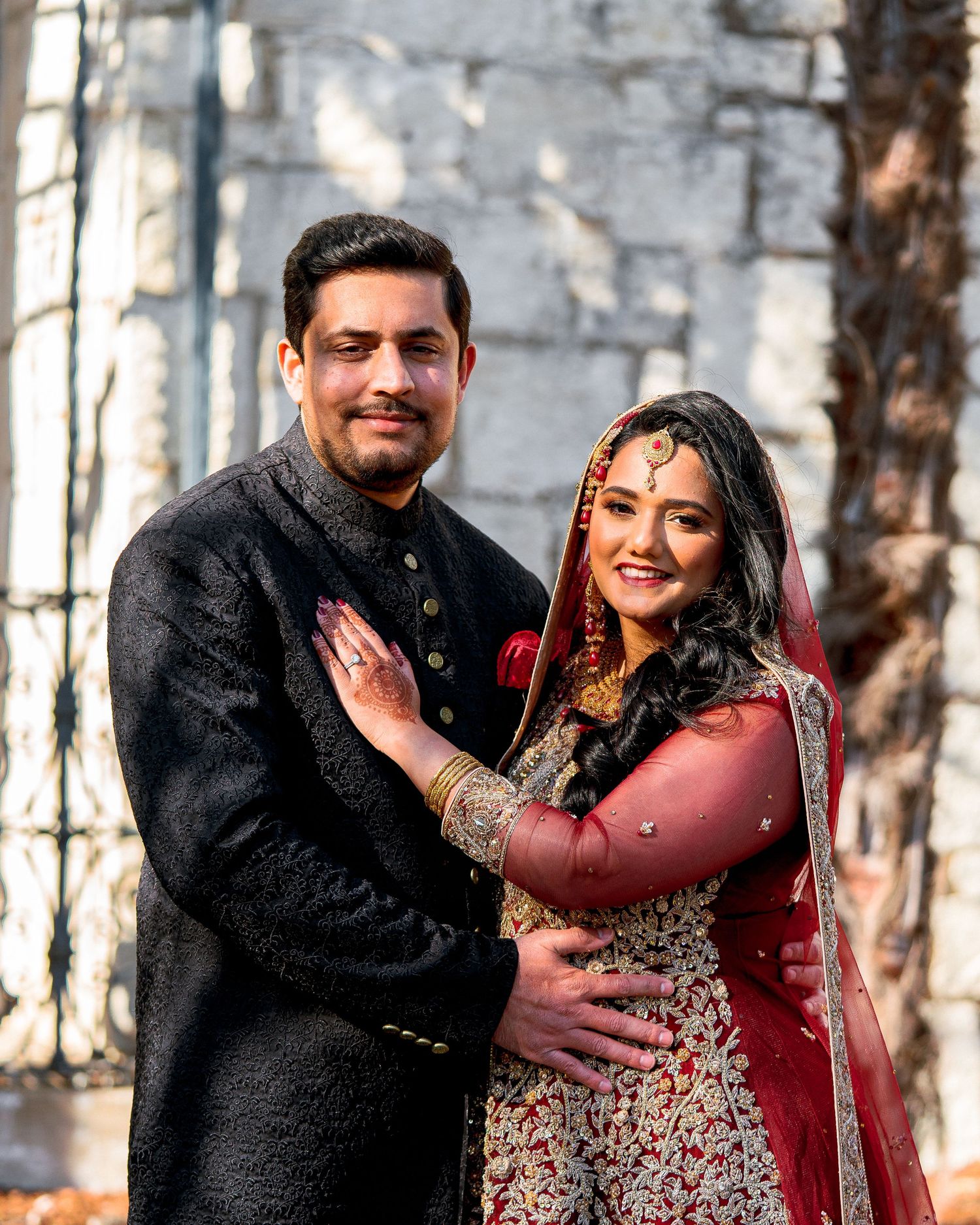 A couple in traditional South Asian wedding attire embraces while laughing outdoors in a forest setting.