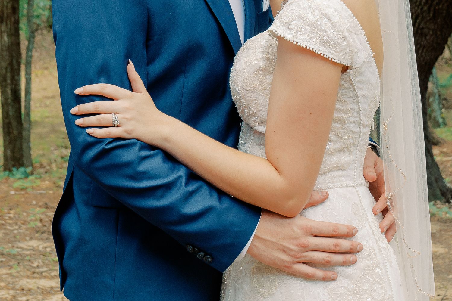 Close-up of bride in lace dress and groom in navy suit embracing during outdoor wedding ceremony.