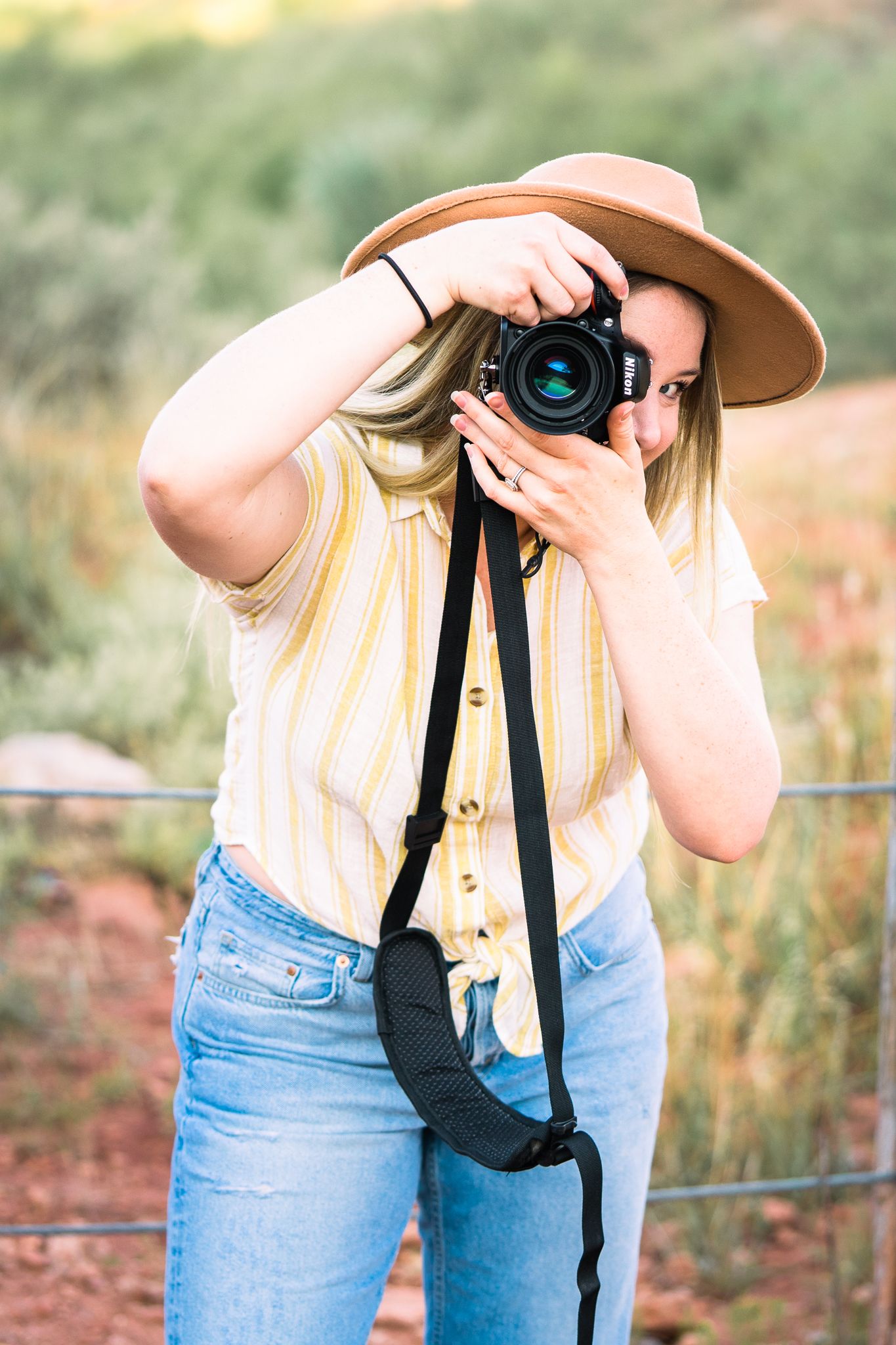 Photographer in yellow shirt and jeans holds camera while standing in outdoor setting during golden hour.