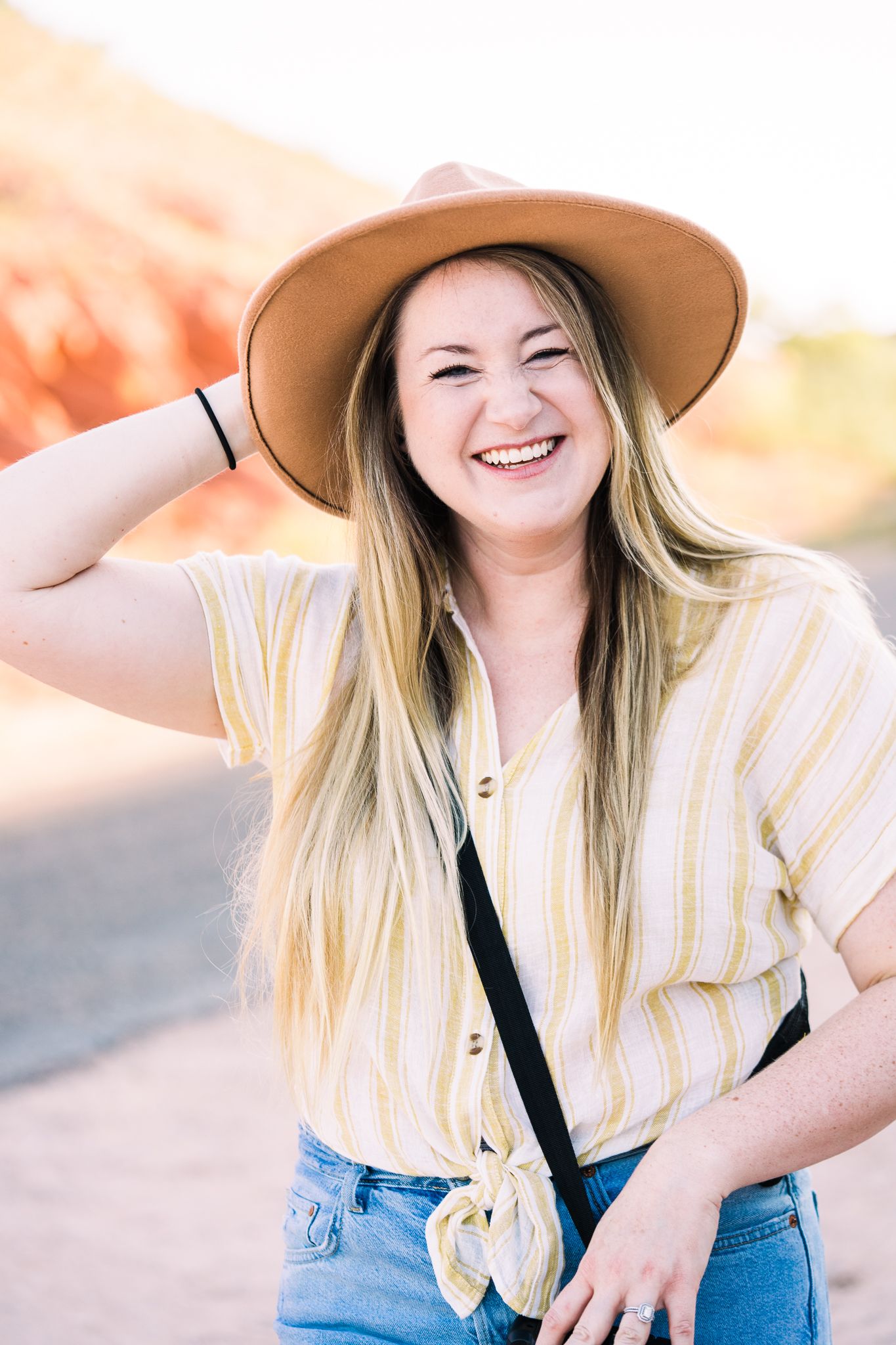 Woman in tan hat and striped shirt laughing joyfully outdoors during sunset.
