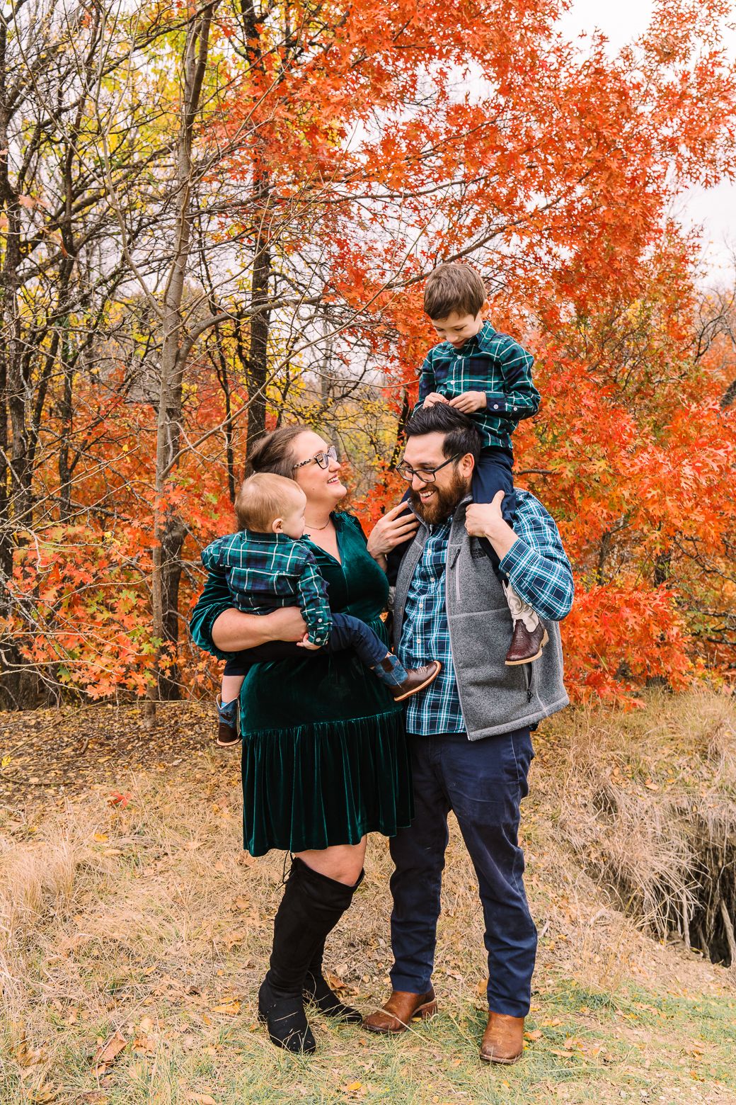 Family wearing plaid embraces in a scenic autumn setting with vibrant orange and yellow foliage in the background.