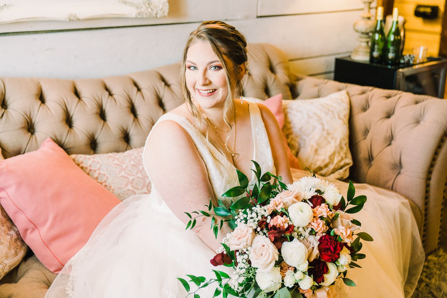 A bridal bouquet of white and burgundy flowers rests on a beige tufted couch with pink accent pillows.