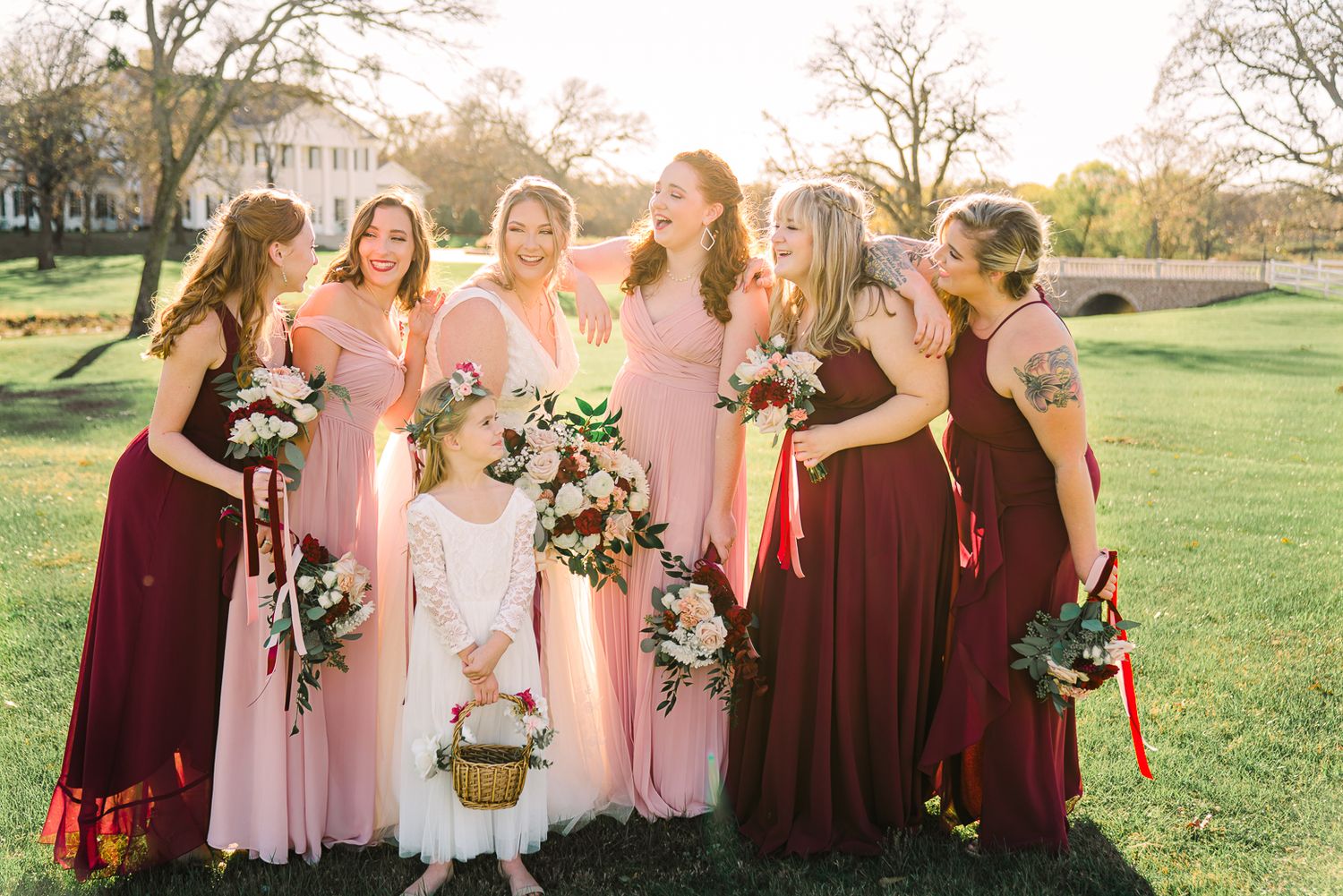 A group in pink and burgundy dresses stands together holding floral bouquets in a sunlit outdoor setting.