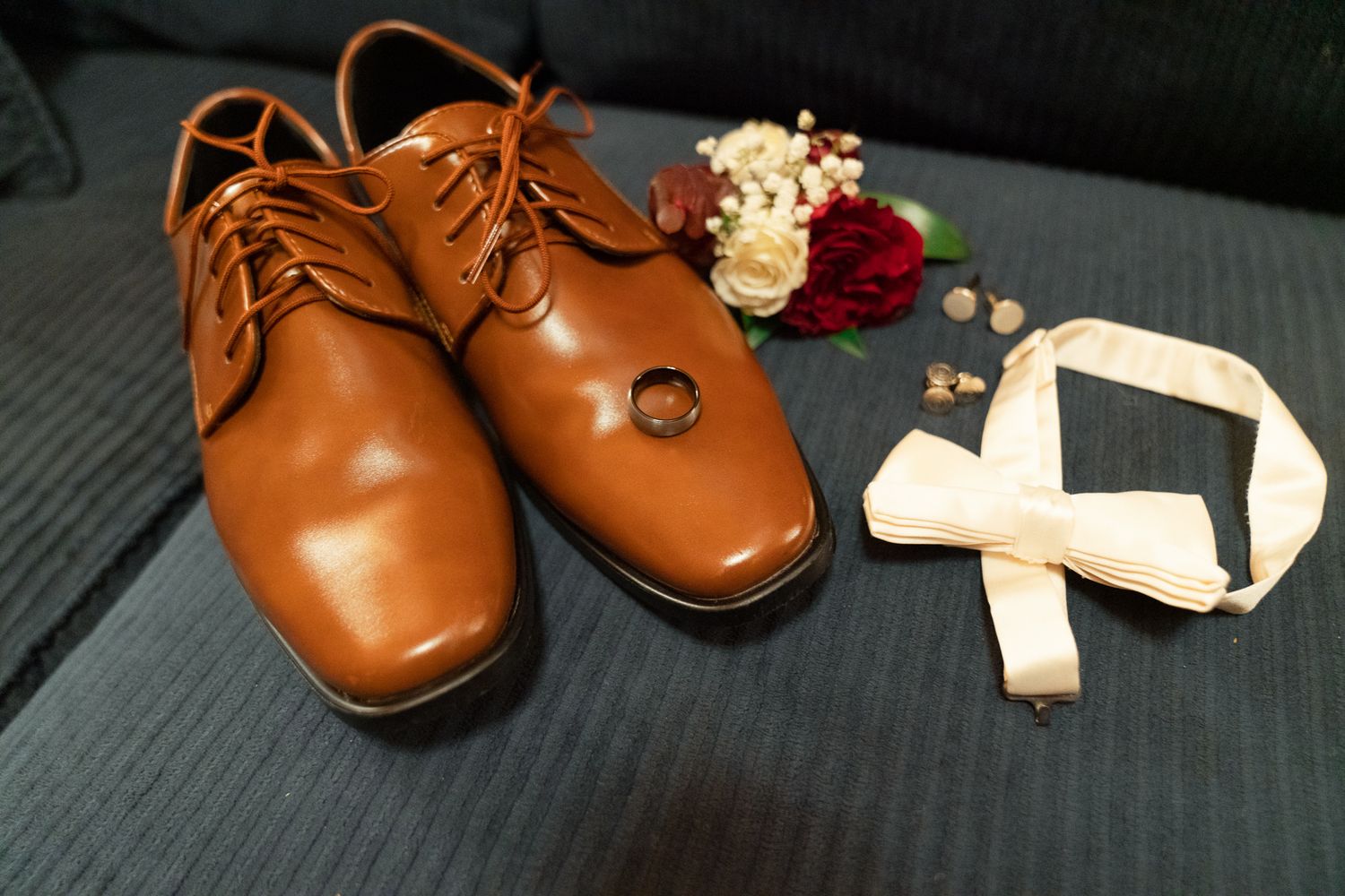 Brown leather dress shoes with ribbons and floral accessories arranged on dark surface.