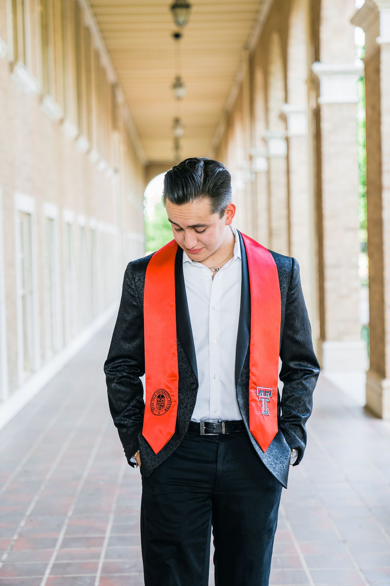 A graduate wearing a black suit and red graduation stole stands thoughtfully in an elegant covered walkway.