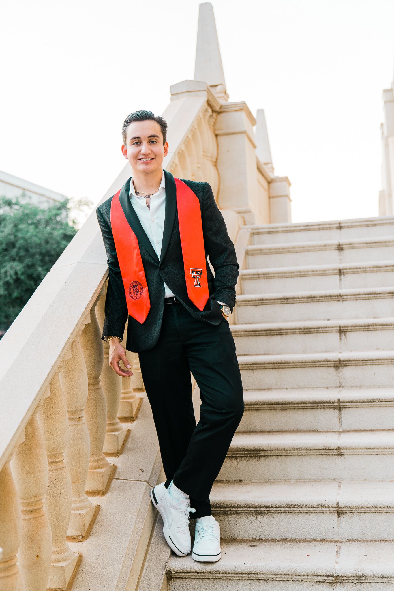 Graduate wearing black suit with red stole poses on ornate stone steps during graduation photoshoot.