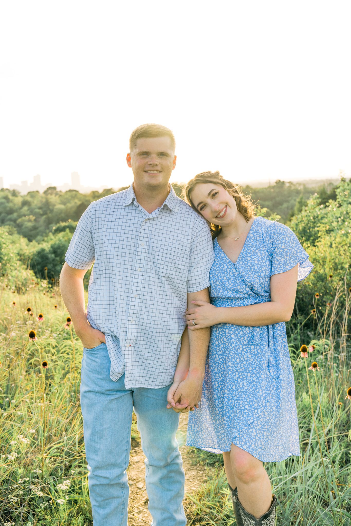 Couple in coordinating blue outfits embrace in a sunlit wildflower field during golden hour.