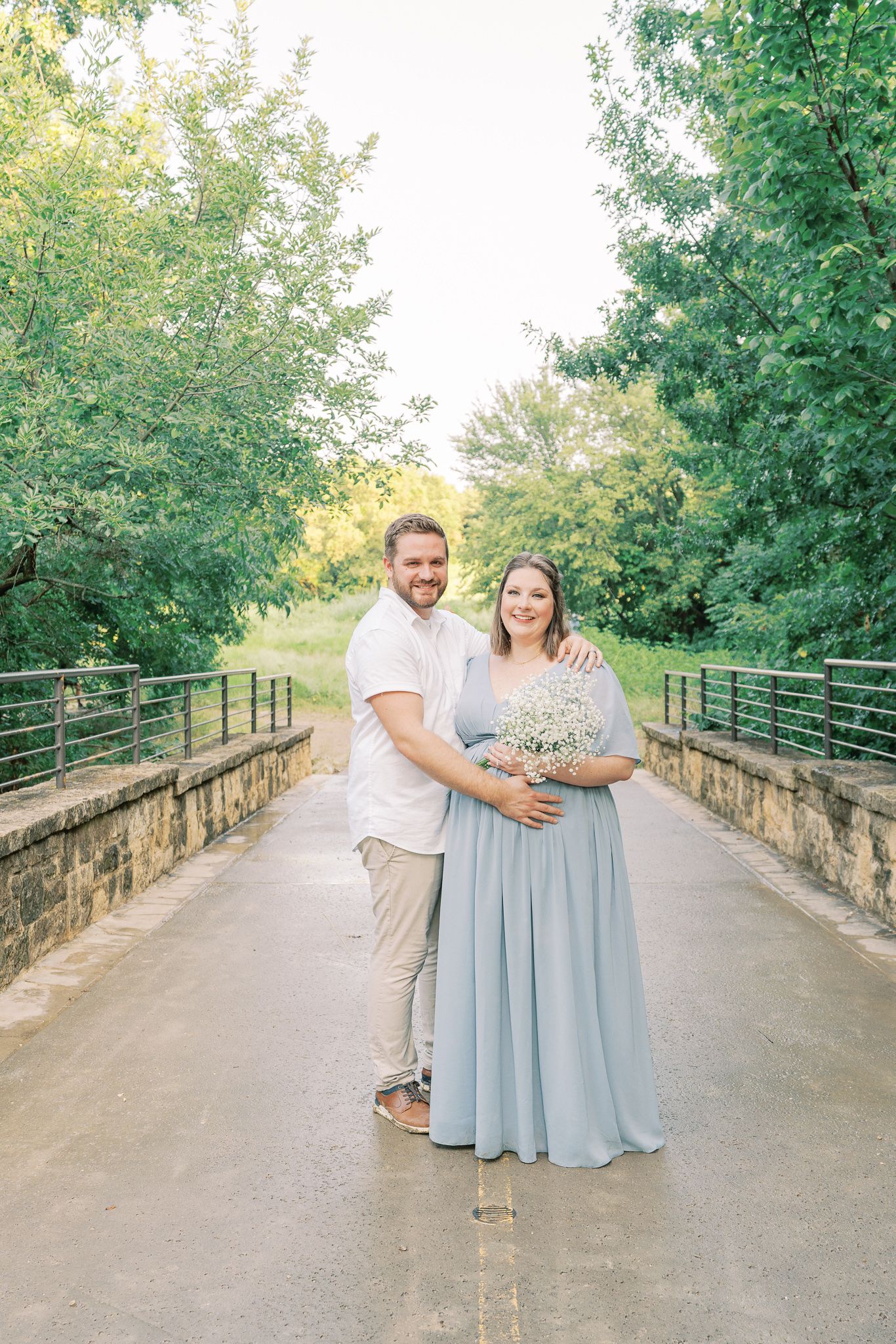A couple poses together on a stone bridge surrounded by lush green trees on a sunny day.